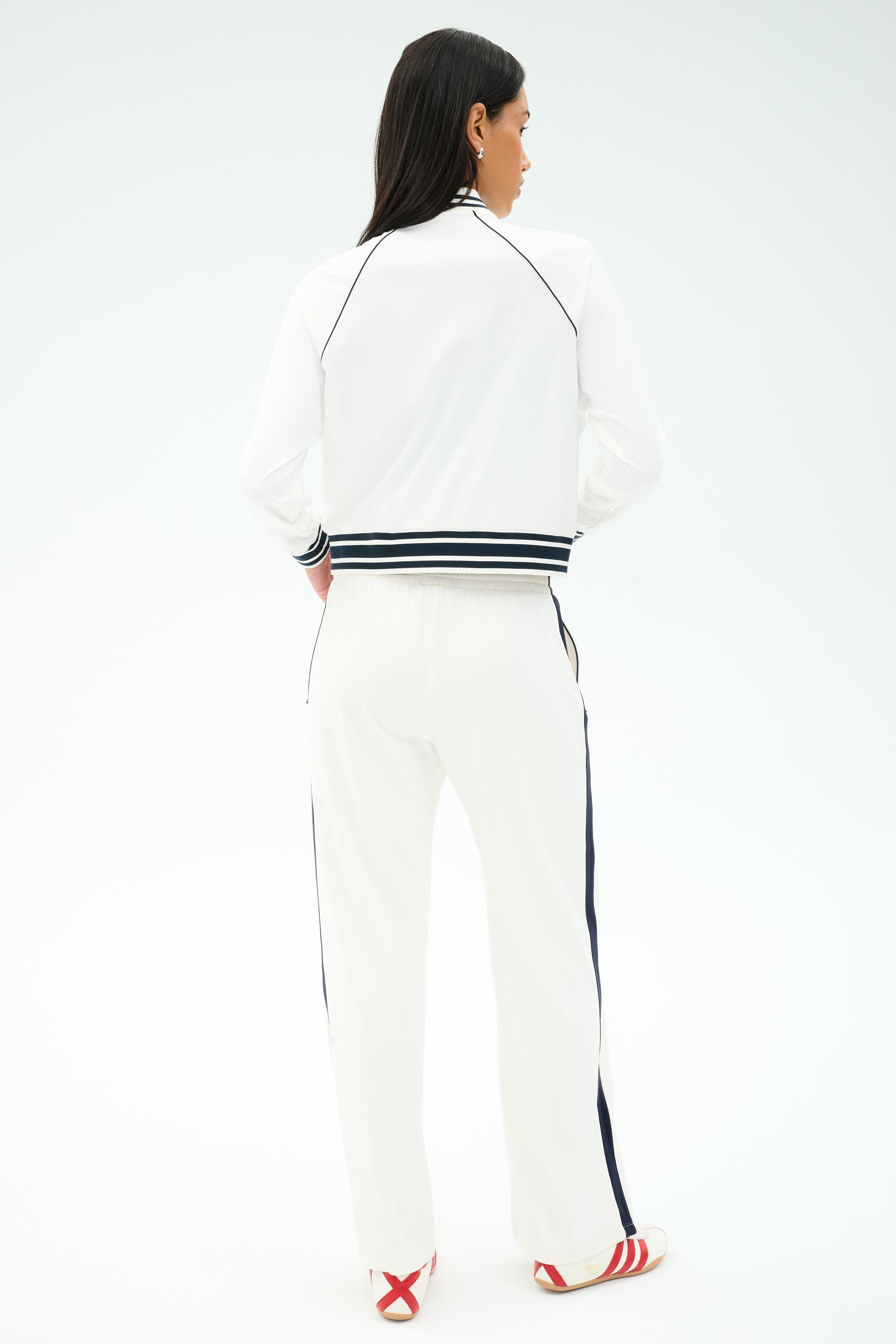 A person stands facing away in the SPLITS59 Hayes Varsity Rigor Jacket in white, paired with black-trimmed pants and red-and-white sandals, set against a plain light background.