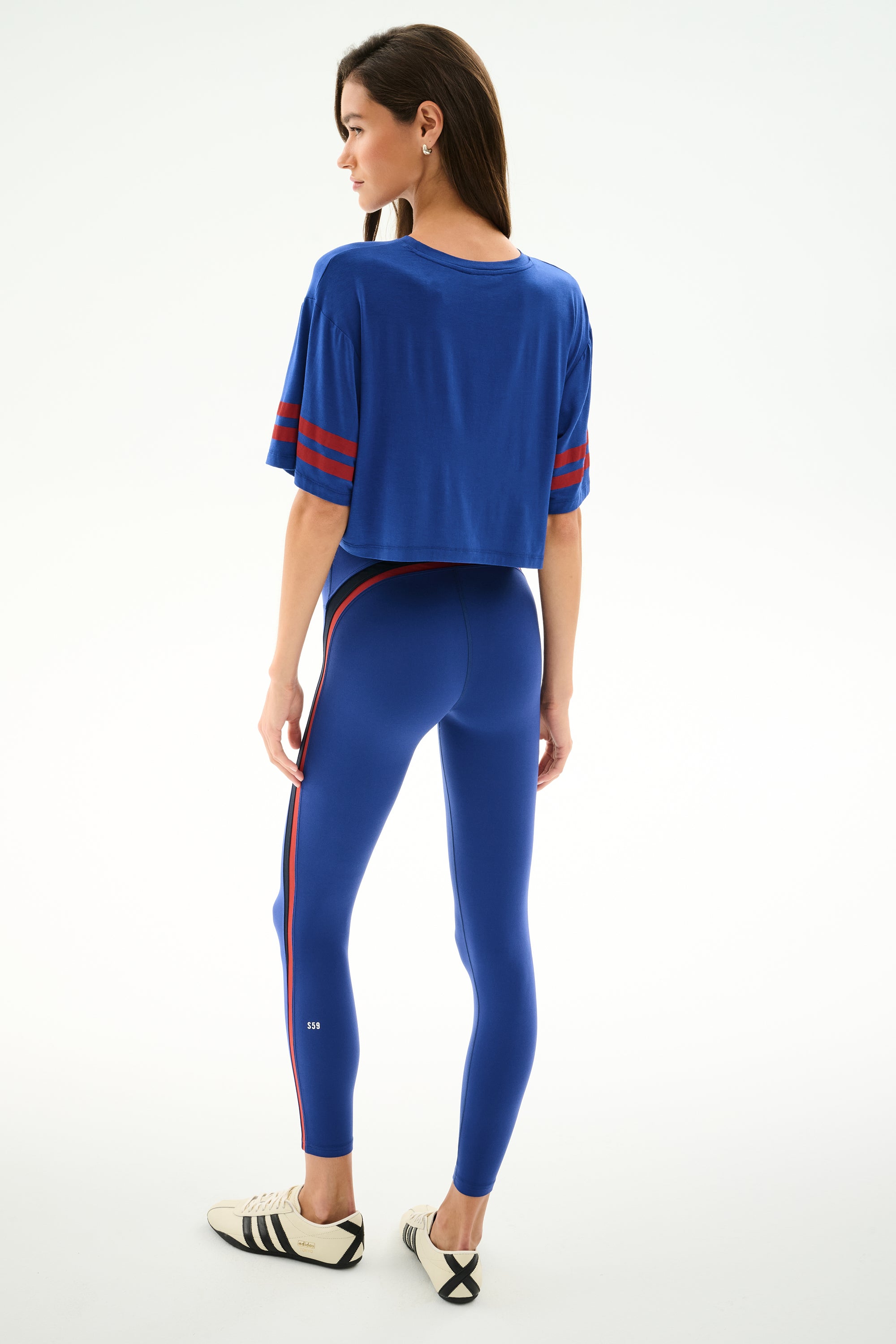 Woman facing away in SPLITS59 Djuna Oversized Crop Jersey Tee - Cobalt/Terracotta and blue leggings, with white sneakers, on a plain white background.