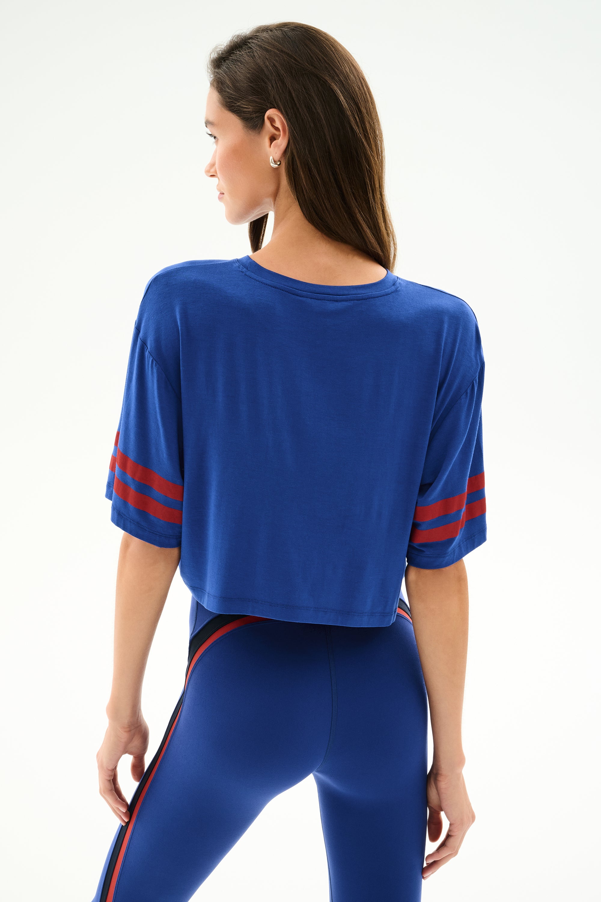 A woman faces away in the Djuna Oversized Crop Jersey Tee - Cobalt/Terracotta by SPLITS59, paired with high-waisted spandex leggings, against a plain white background.