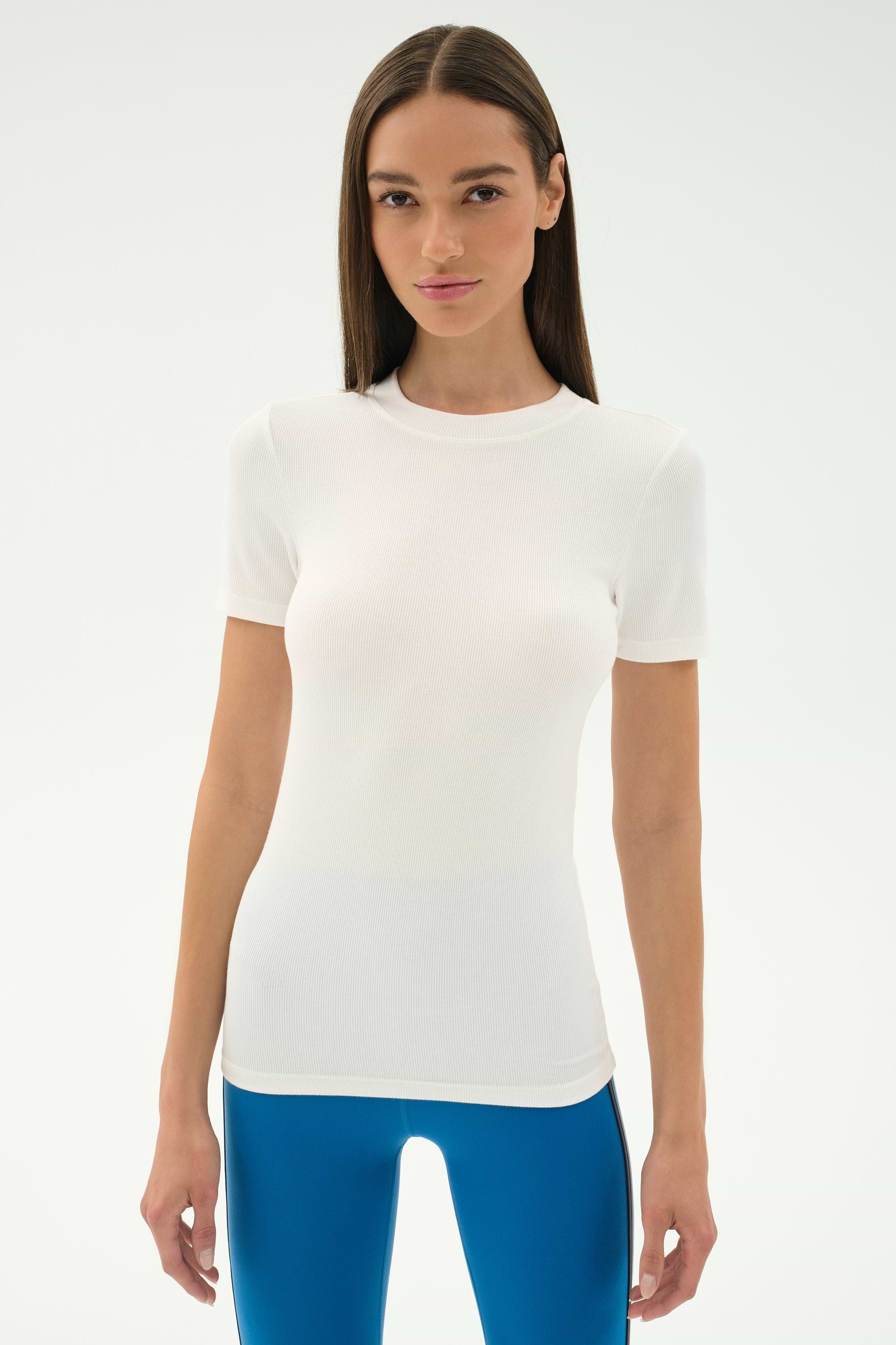 A woman with straight brown hair wears the SPLITS59 Louise Rib Short Sleeve in white and shiny blue leggings, standing against a plain light background.