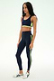 A woman is wearing the SPLITS59 Maci Airweight Bra in Indigo/Limeade, featuring neon yellow-green accents and crafted from a stretchy nylon spandex fabric, paired with white sneakers against a plain light background.