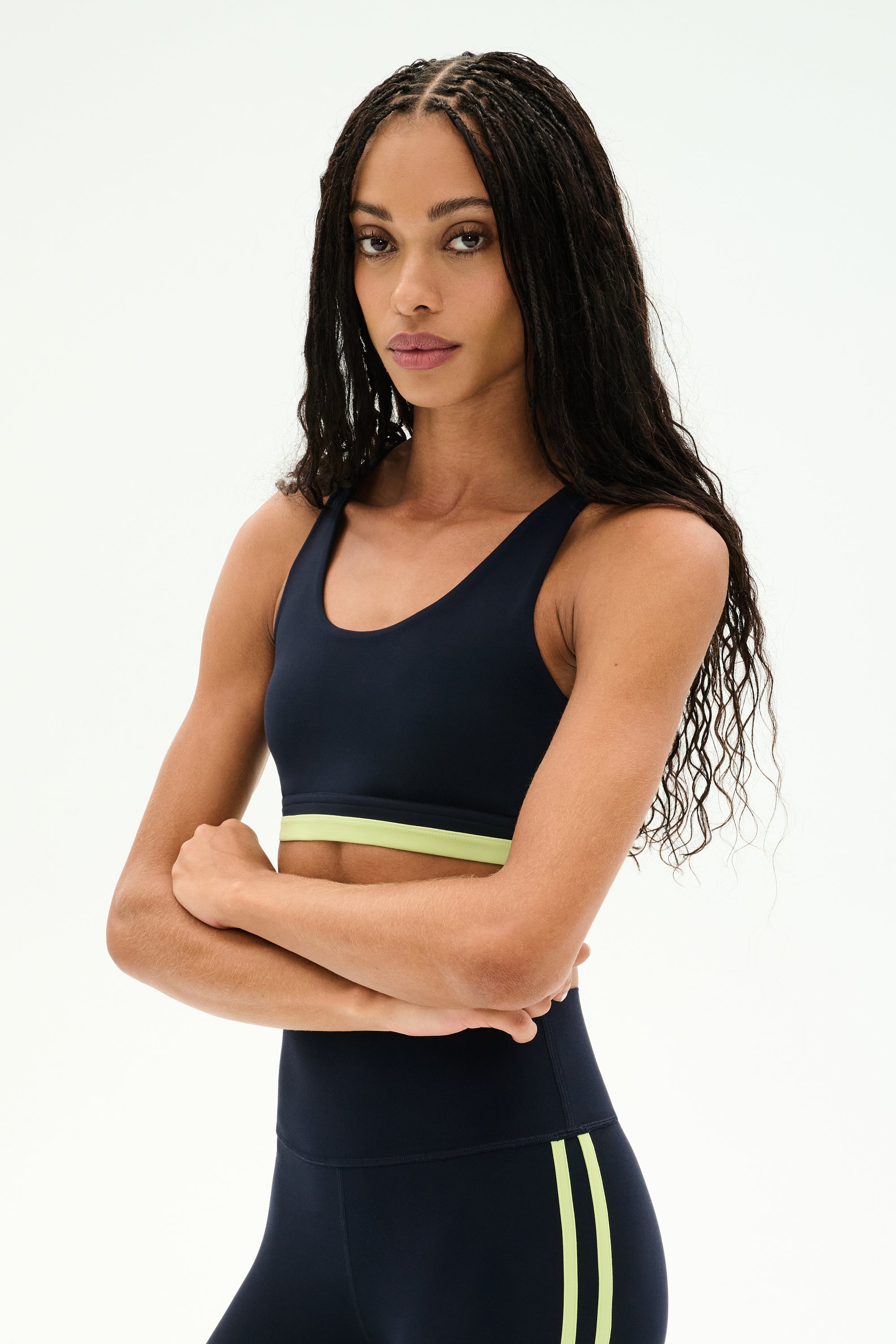 A woman with long braided hair stands against a white background, arms crossed, wearing the SPLITS59 Maci Airweight Bra in Indigo/Limeade—a navy and lime green activewear piece made from stretchy nylon spandex fabric.