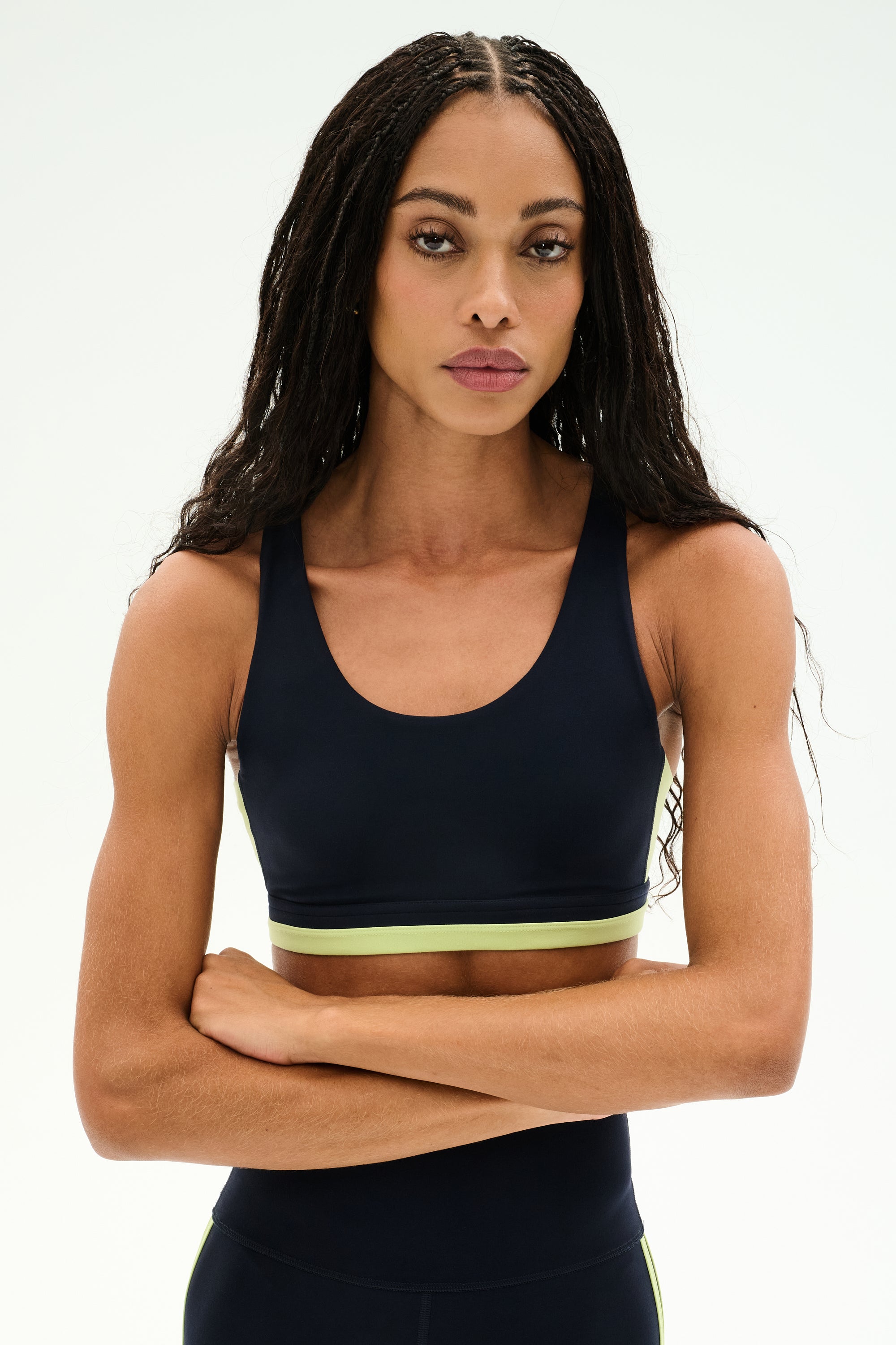 A woman with long braided hair stands with her arms crossed against a plain light background, wearing the SPLITS59 Maci Airweight Bra in Indigo/Limeade, made from stretchy nylon-spandex activewear fabric.