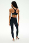 A woman stands facing away, wearing the SPLITS59 Maci Airweight Bra in Indigo/Limeade paired with matching activewear leggings featuring a side stripe, all crafted from stretchy nylon spandex fabric, and white sneakers against a plain white background.