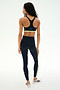 A woman stands facing away, wearing the SPLITS59 Maci Airweight Bra in Indigo/Limeade paired with matching activewear leggings featuring a side stripe, all crafted from stretchy nylon spandex fabric, and white sneakers against a plain white background.
