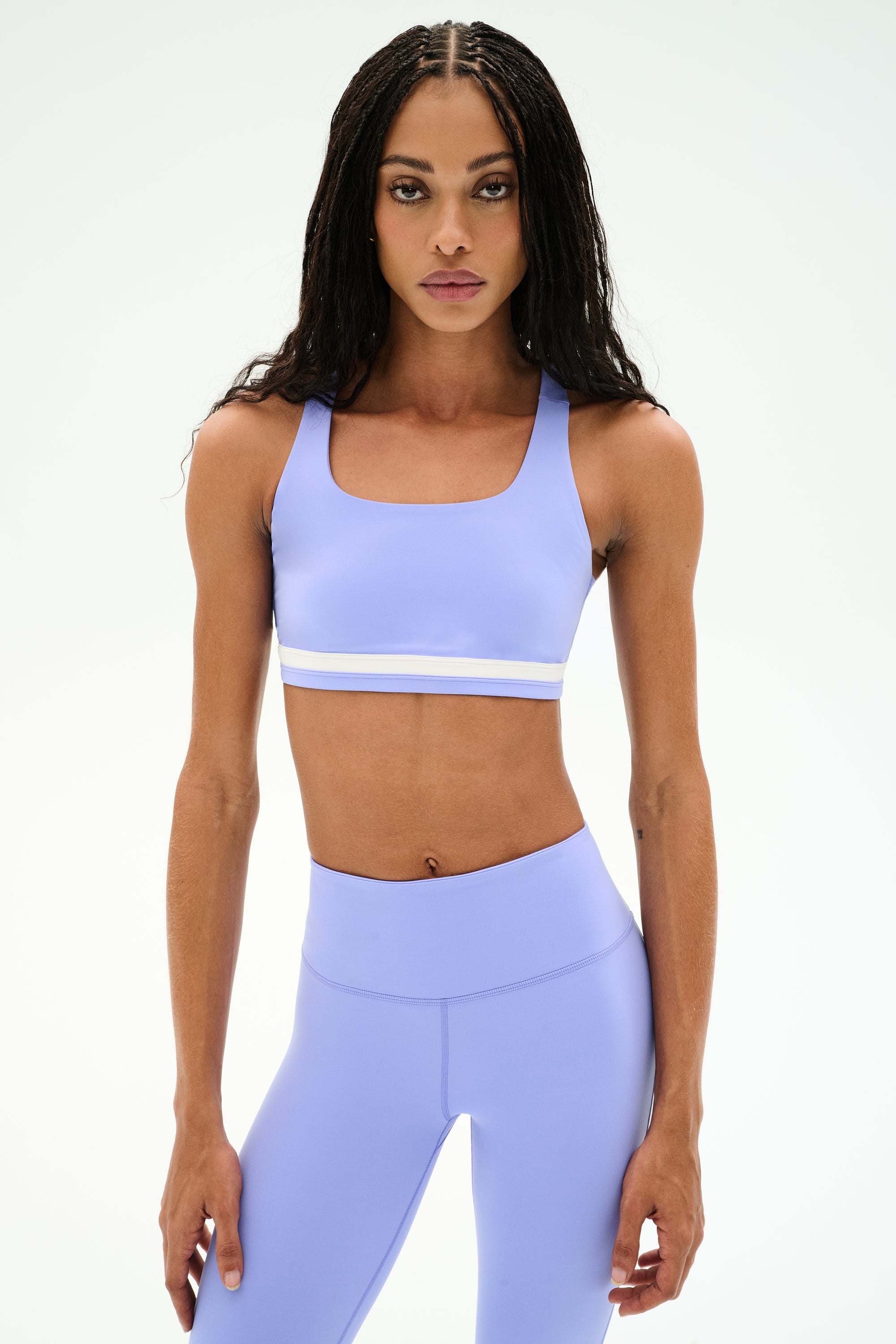 A woman wearing a SPLITS59 Miles Rigor Bra in Purple Haze/White with matching leggings stands facing forward against a plain white background.