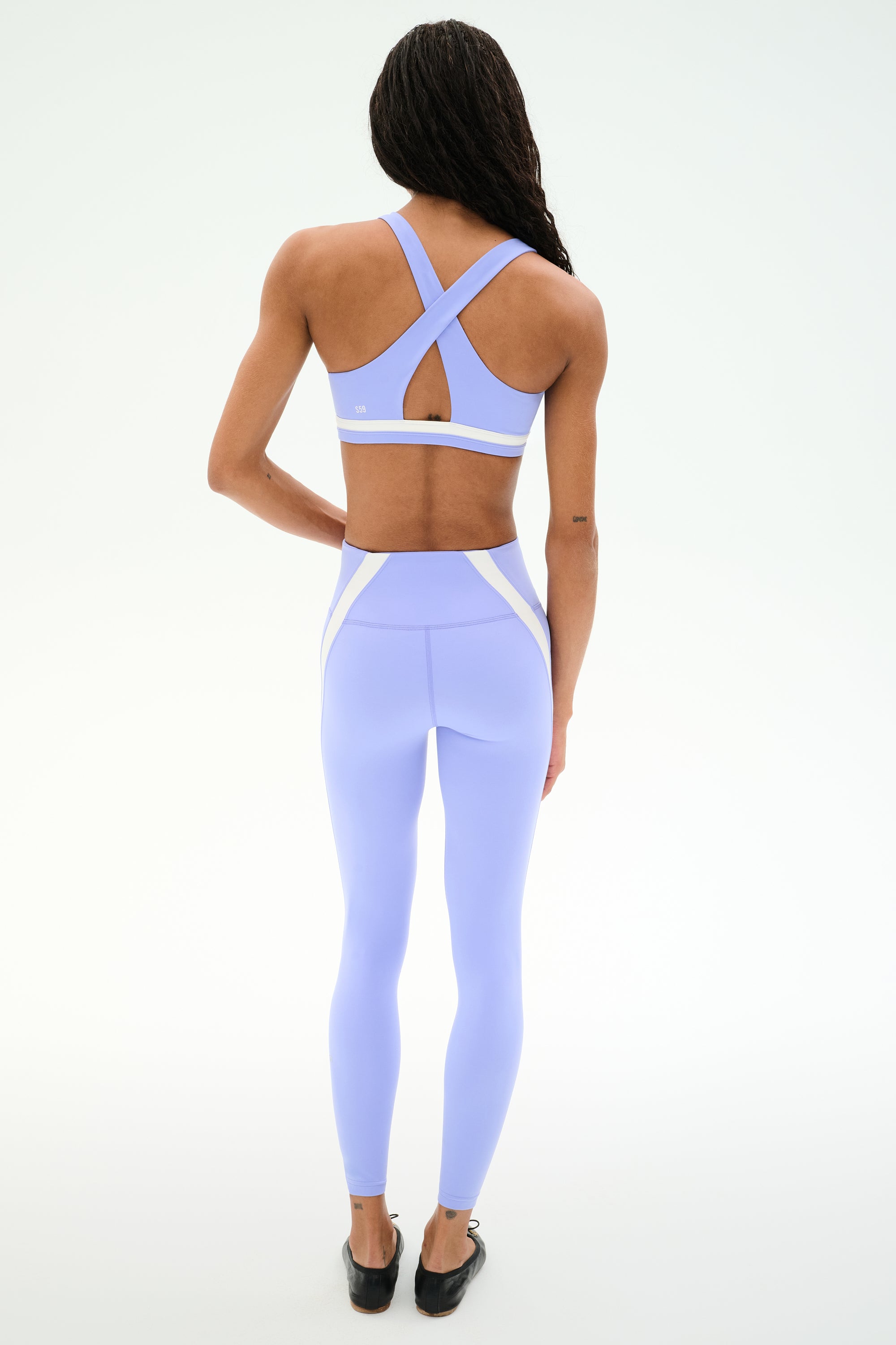 A person faces away, wearing the SPLITS59 Miles Rigor Bra in Purple Haze/White with crisscross straps and high-waisted leggings, set against a plain white background.