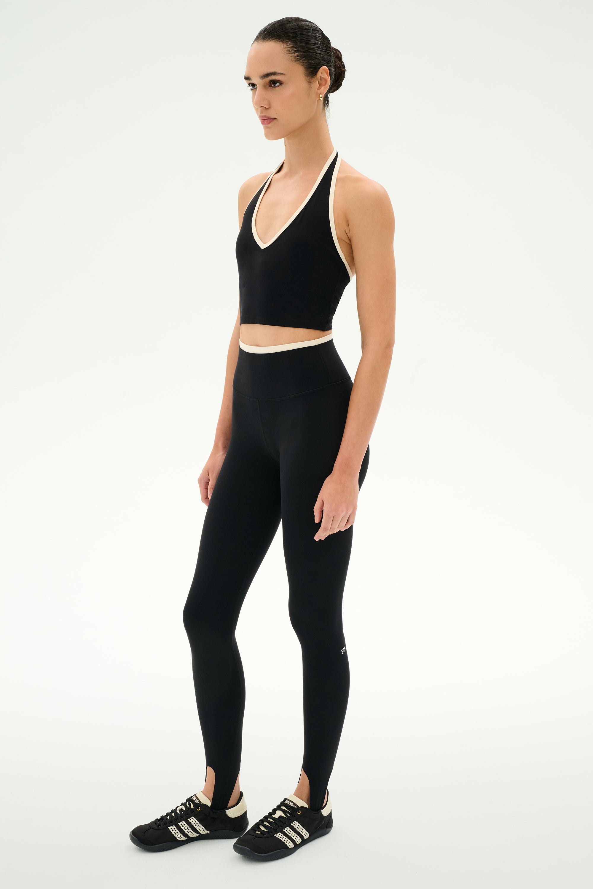 A woman stands in a studio, facing slightly to the side against a plain white background, wearing the SPLITS59 Rory Airweight Halter Bralette in Black/Creme, paired with high-waisted leggings and black sneakers.