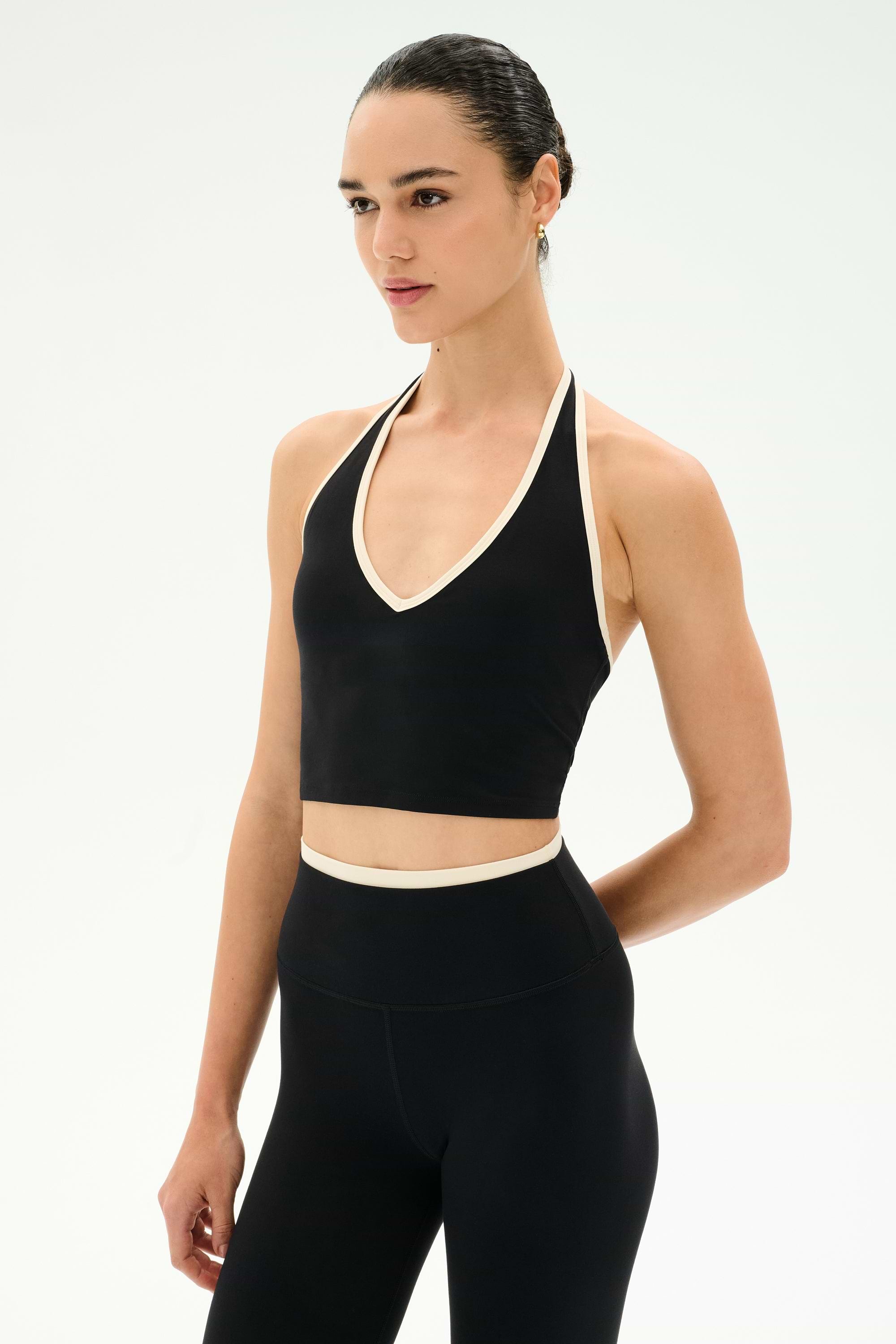 A woman models the SPLITS59 Rory Airweight Halter Bralette in Black/Creme, a black sleeveless crop top with white trim, crafted from a soft blend of nylon and spandex, shown against a plain white background.