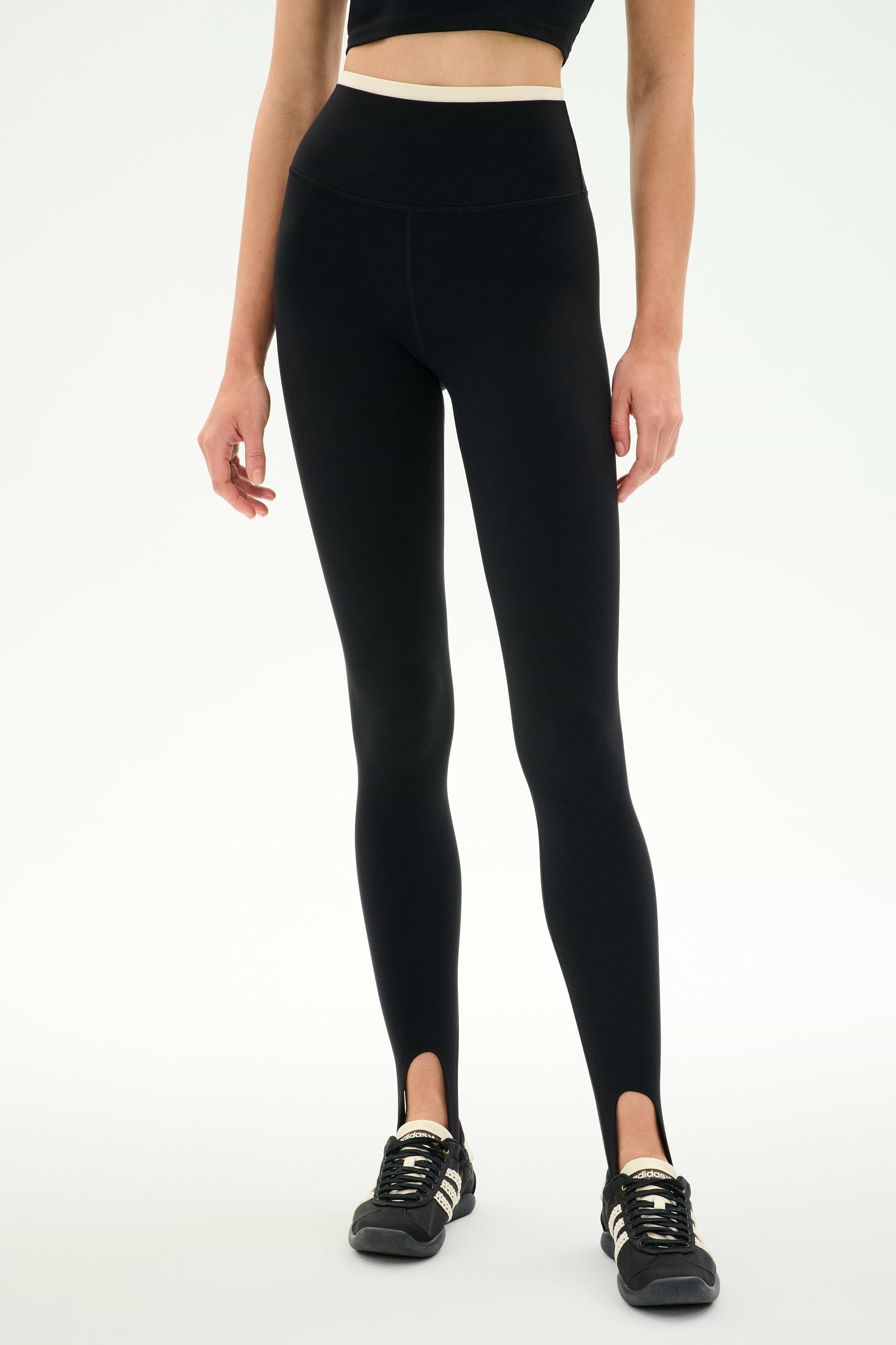 A woman stands against a plain white background wearing the SPLITS59 River Dual Waist Airweight Stirrup leggings in Black/Creme, crafted from a stretchy nylon spandex athletic fabric, paired with black sneakers. Her face is not visible.
