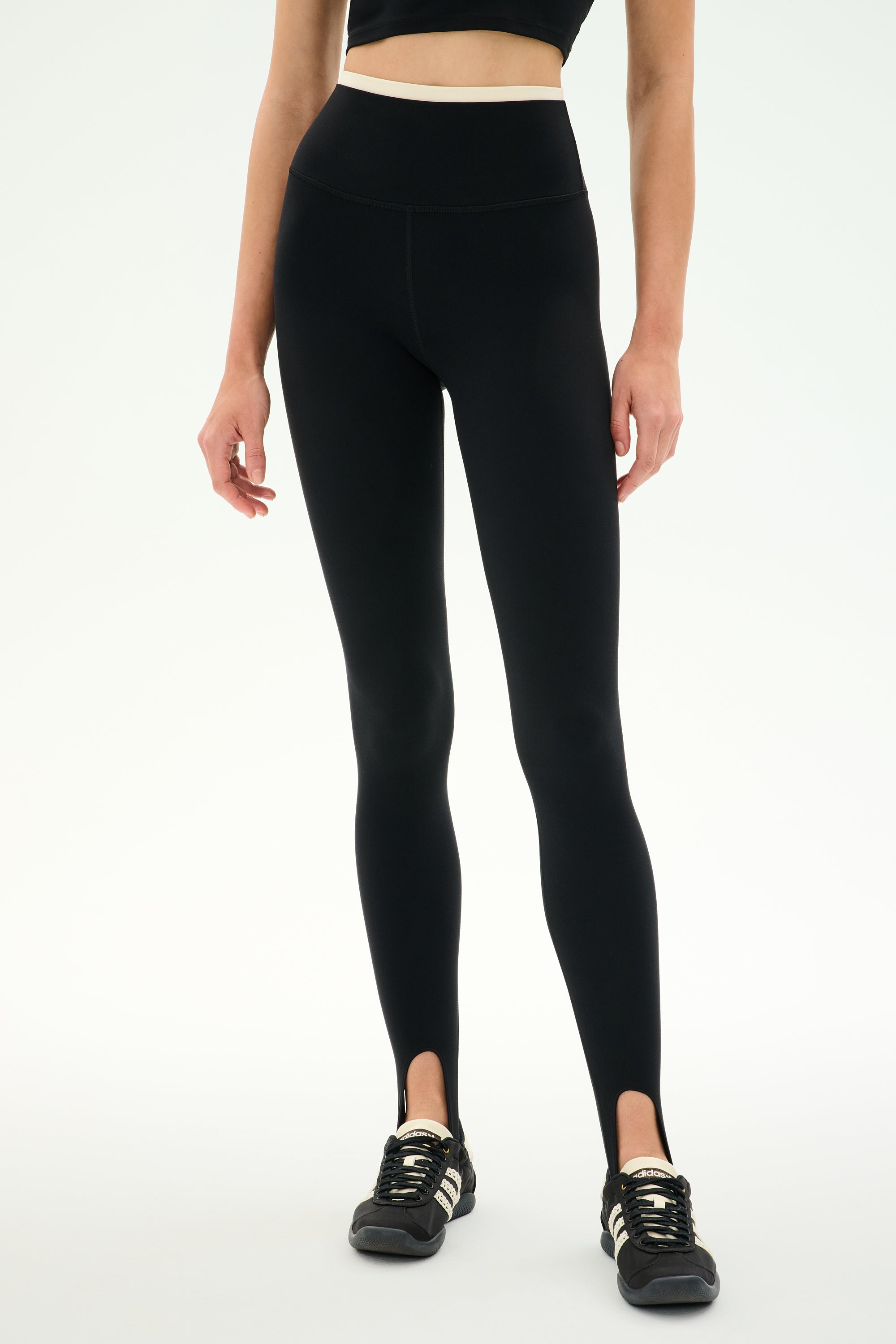 A woman stands against a plain white background wearing the SPLITS59 River Dual Waist Airweight Stirrup leggings in Black/Creme, crafted from a stretchy nylon spandex athletic fabric, paired with black sneakers. Her face is not visible.