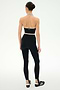 A woman stands facing away, wearing the SPLITS59 Rory Airweight Halter Bralette in Black/Creme and high-waisted black leggings crafted from a sleek nylon-spandex blend, along with black athletic shoes featuring white stripes, all set against a plain white background.