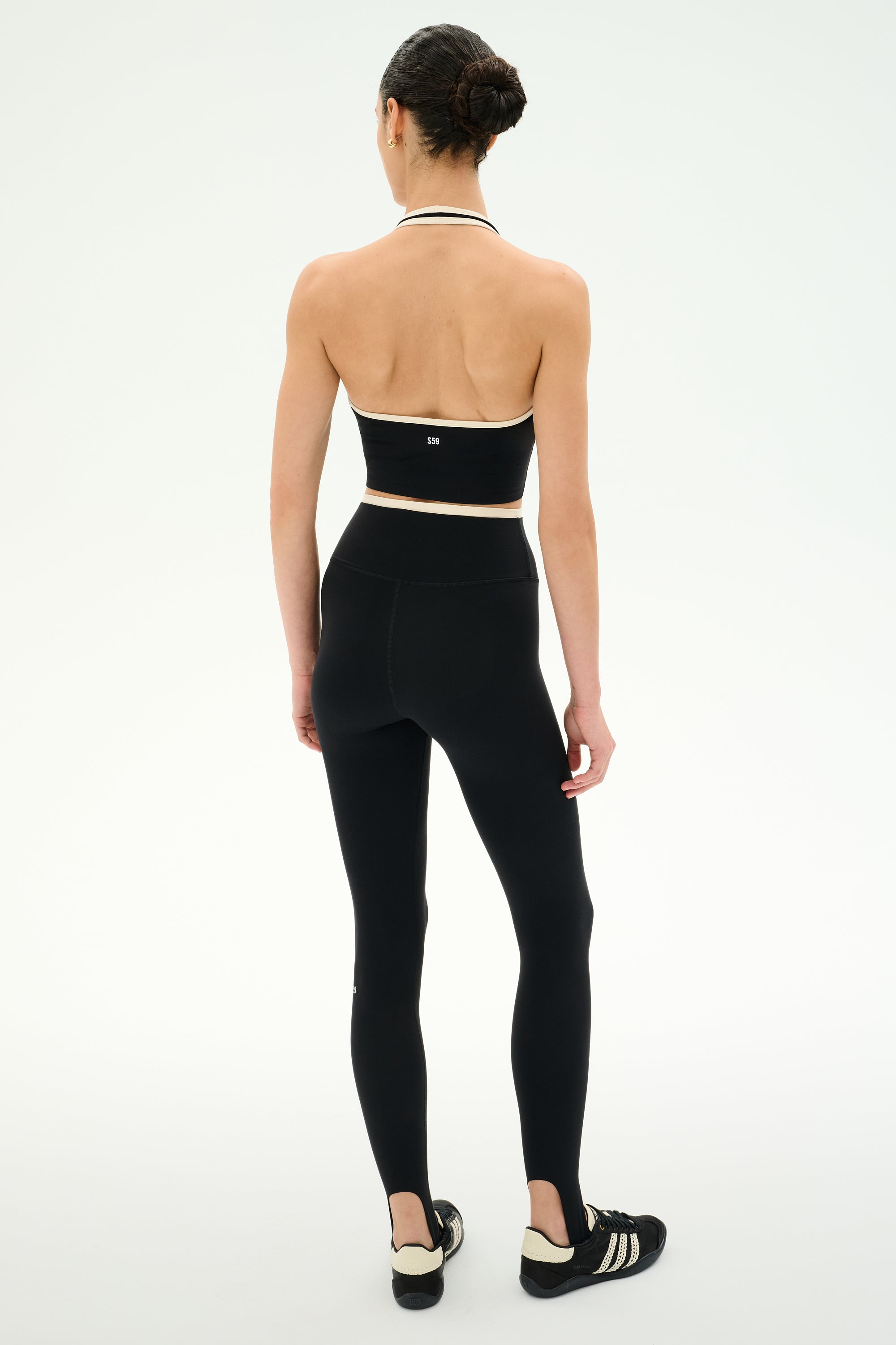 A woman stands facing away, wearing the SPLITS59 Rory Airweight Halter Bralette in Black/Creme and high-waisted black leggings crafted from a sleek nylon-spandex blend, along with black athletic shoes featuring white stripes, all set against a plain white background.