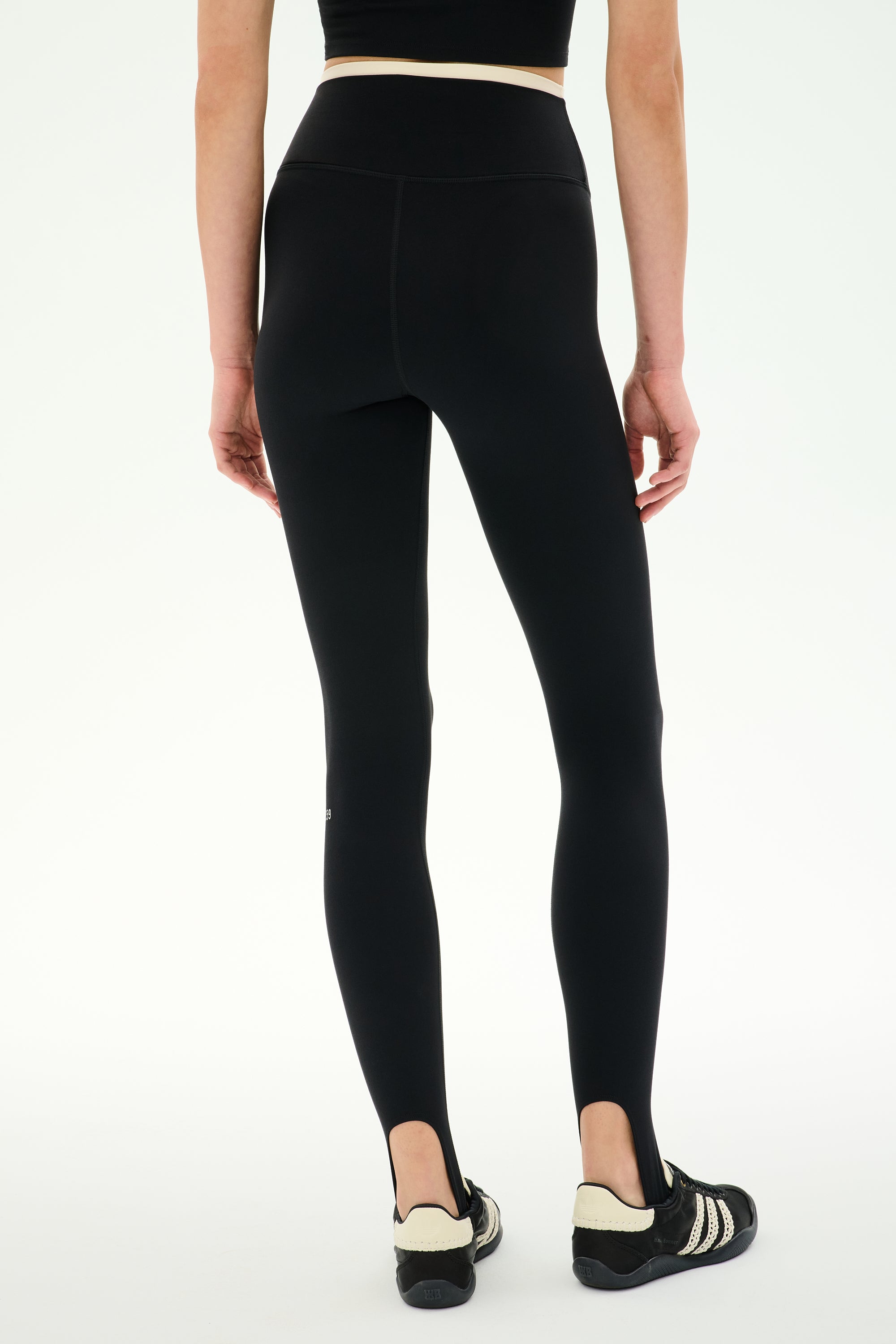 Model wearing the SPLITS59 River Dual Waist Airweight Stirrup - Black/Creme leggings with foot straps, crafted from stretch material, paired with black athletic shoes. Shown from the back against a plain white background.