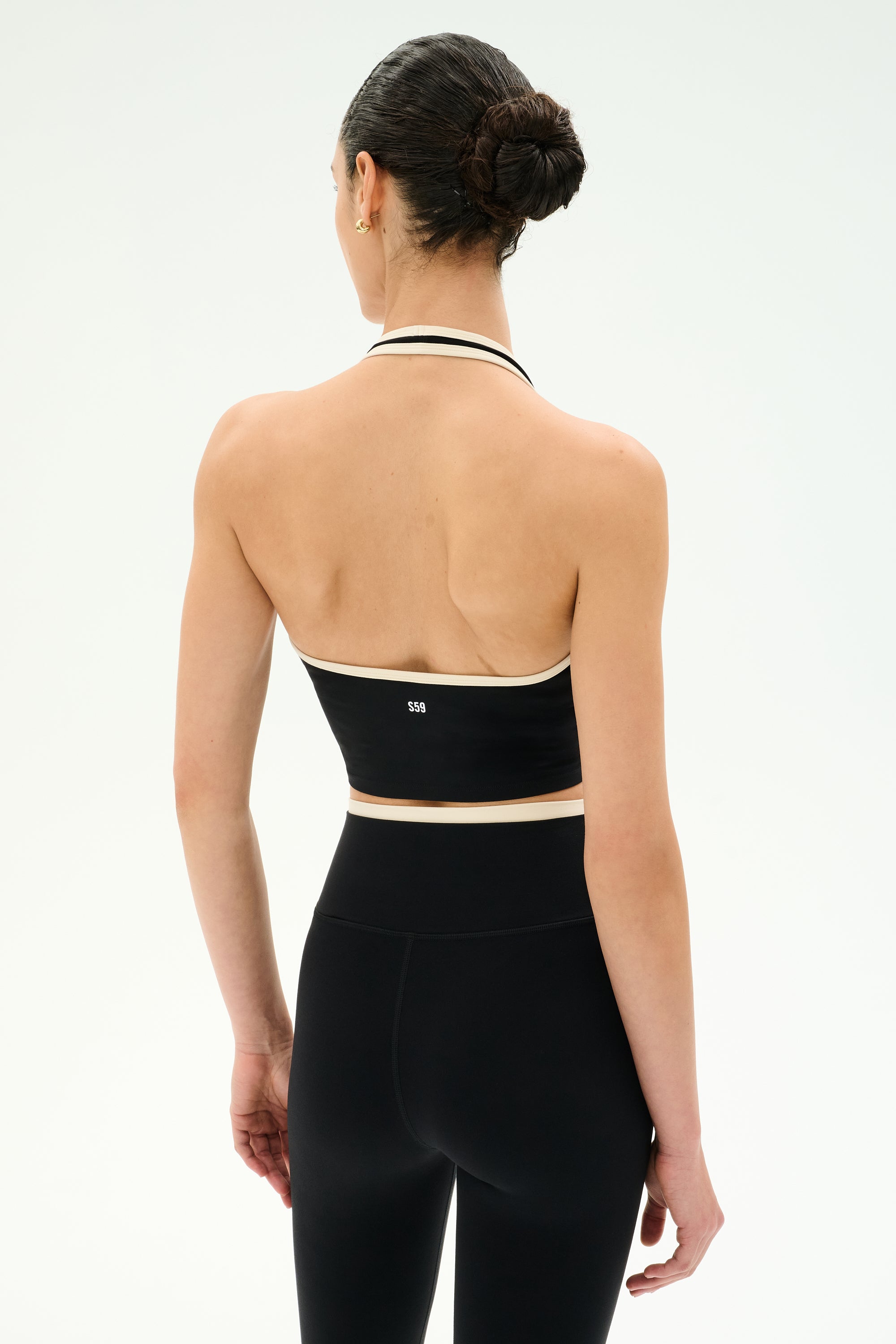 A person with dark hair in a bun is shown from the back, standing against a plain white background, wearing the SPLITS59 Rory Airweight Halter Bralette in Black/Creme paired with high-waisted spandex leggings.