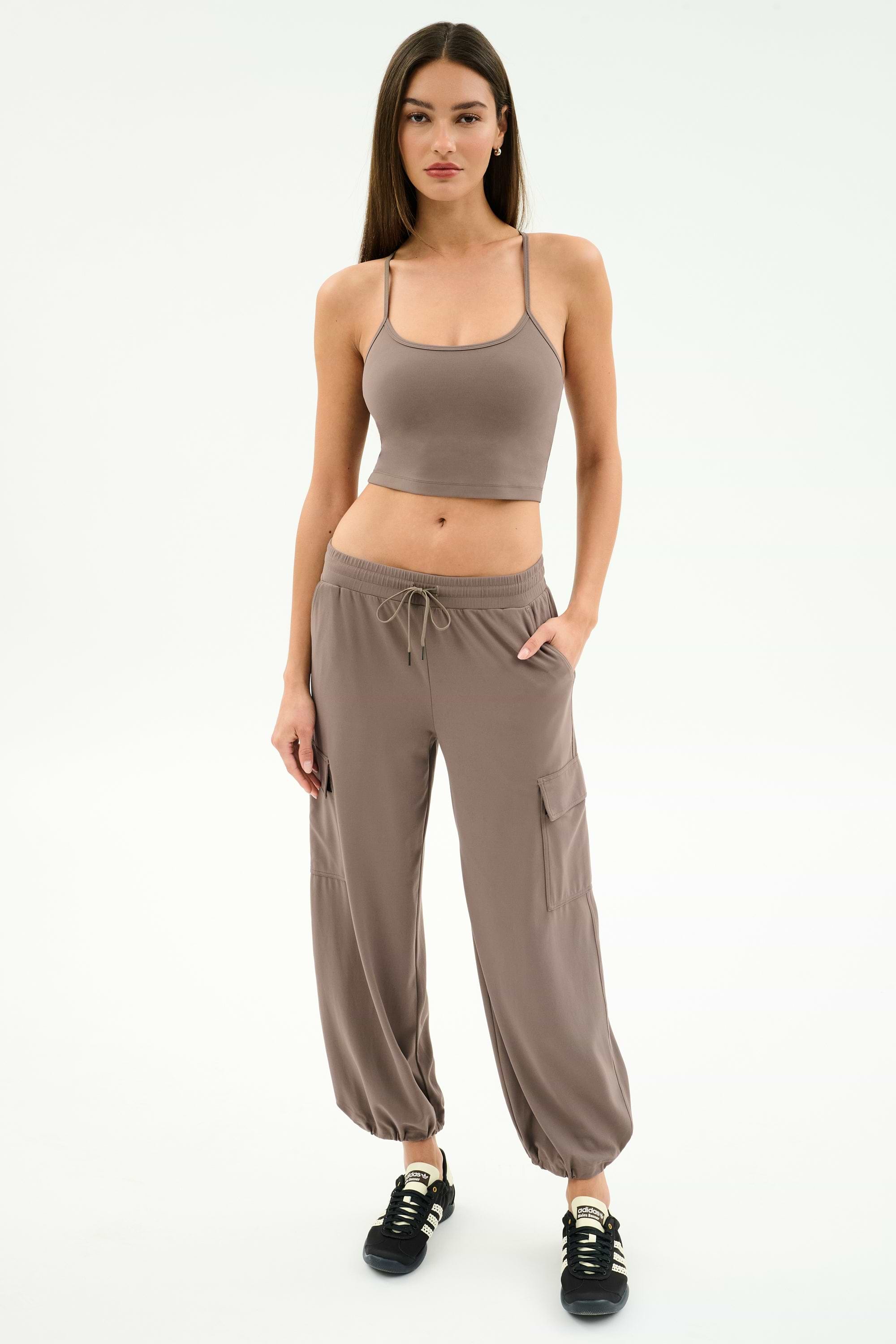 A woman stands against a white background, wearing SPLITS59's Nico Airweight Cargo Pant in Lentil with pockets, styled like joggers, a taupe crop top, and black sneakers.