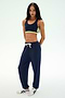 Standing against a plain background, this person is wearing a navy sports bra with green trim, the Andie Oversized Fleece Sweatpant in Indigo by SPLITS59—made in Los Angeles—and white sneakers with black stripes, making it a perfect outfit for cool weather workouts.