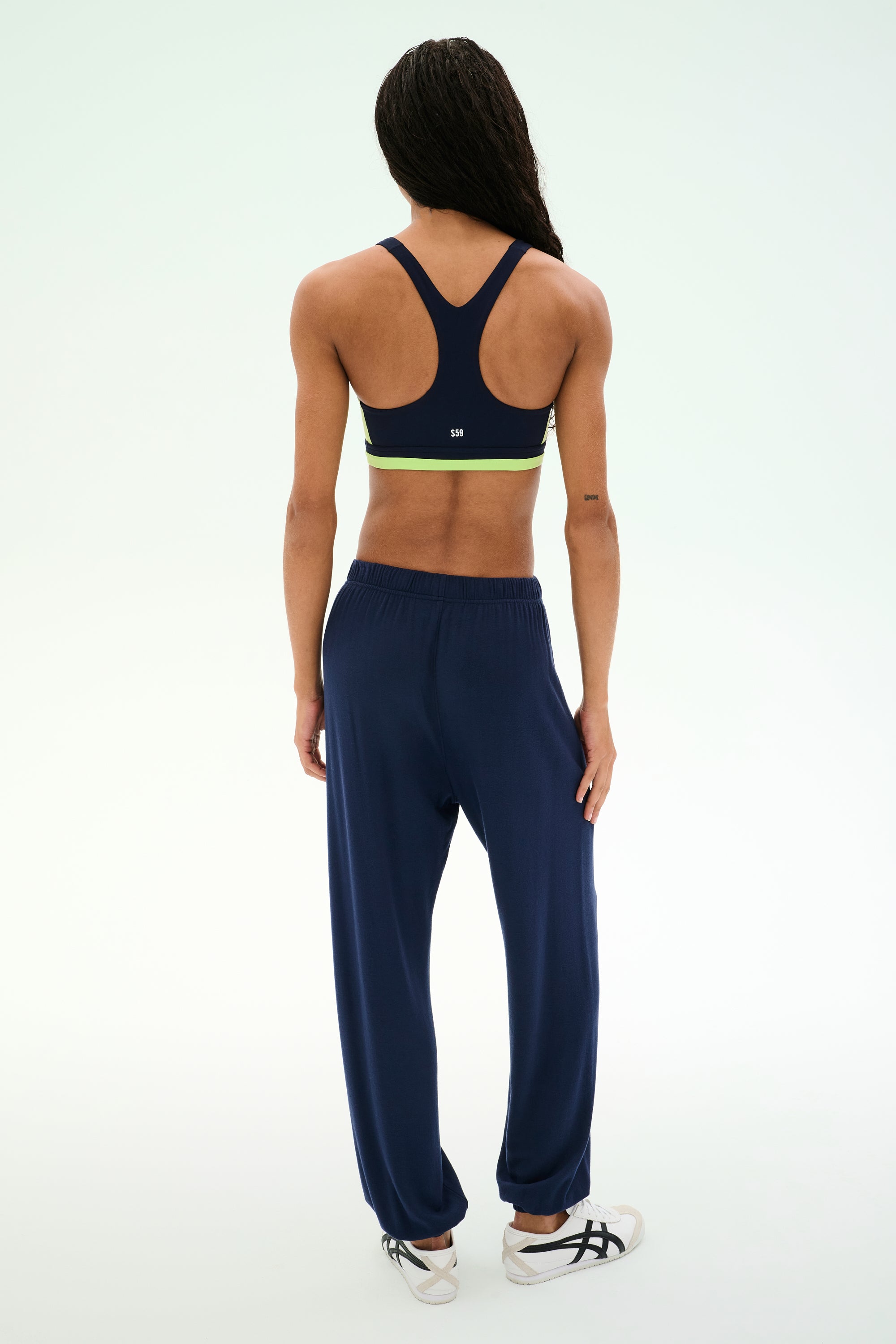 A person stands facing away against a plain light background, wearing a navy sports bra with green trim, white athletic shoes, and the SPLITS59 Andie Oversized Fleece Sweatpant in Indigo—an ideal look for cool weather workouts.