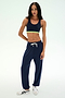 A woman stands against a plain light background in athletic wear—featuring a navy sports bra with green trim, white sneakers, and the Andie Oversized Fleece Sweatpant in Indigo by SPLITS59, made in Los Angeles—perfect for cool weather workouts.