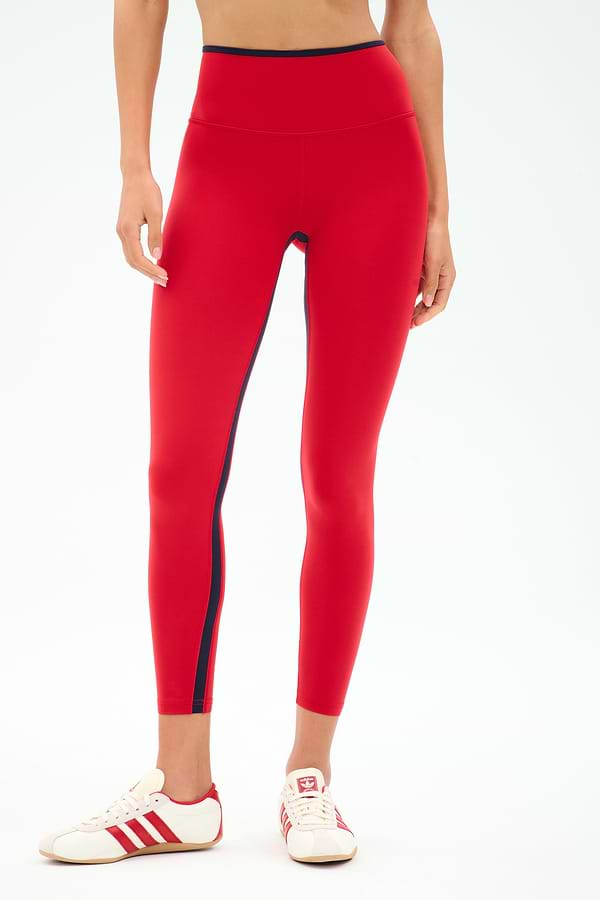 A person wears SPLITS59 Easton Rigor High Waist 7/8 leggings in Pirate Red/Indigo, made from compressive fabric, paired with white sneakers featuring red stripes, standing against a plain background.