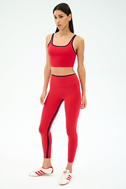 A woman wears the SPLITS59 Easton Rigor High Waist 7/8 in Pirate Red/Indigo, featuring compressive fabric, along with white and red sneakers, standing against a plain light background.