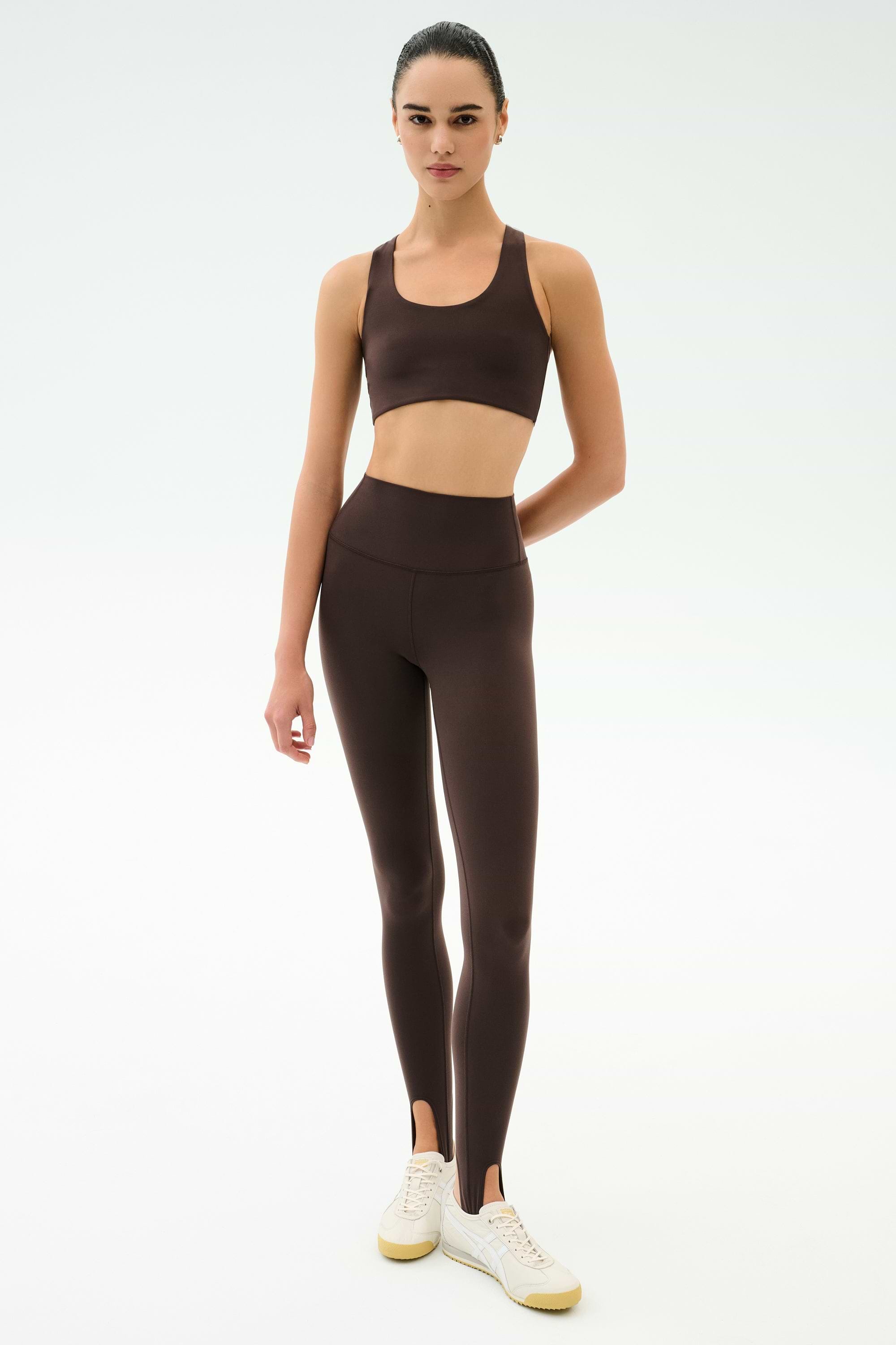 A person stands against a plain background wearing SPLITS59's River High Waist Airweight Stirrup leggings in Dark Chocolate, paired with a matching sports bra and white sneakers.