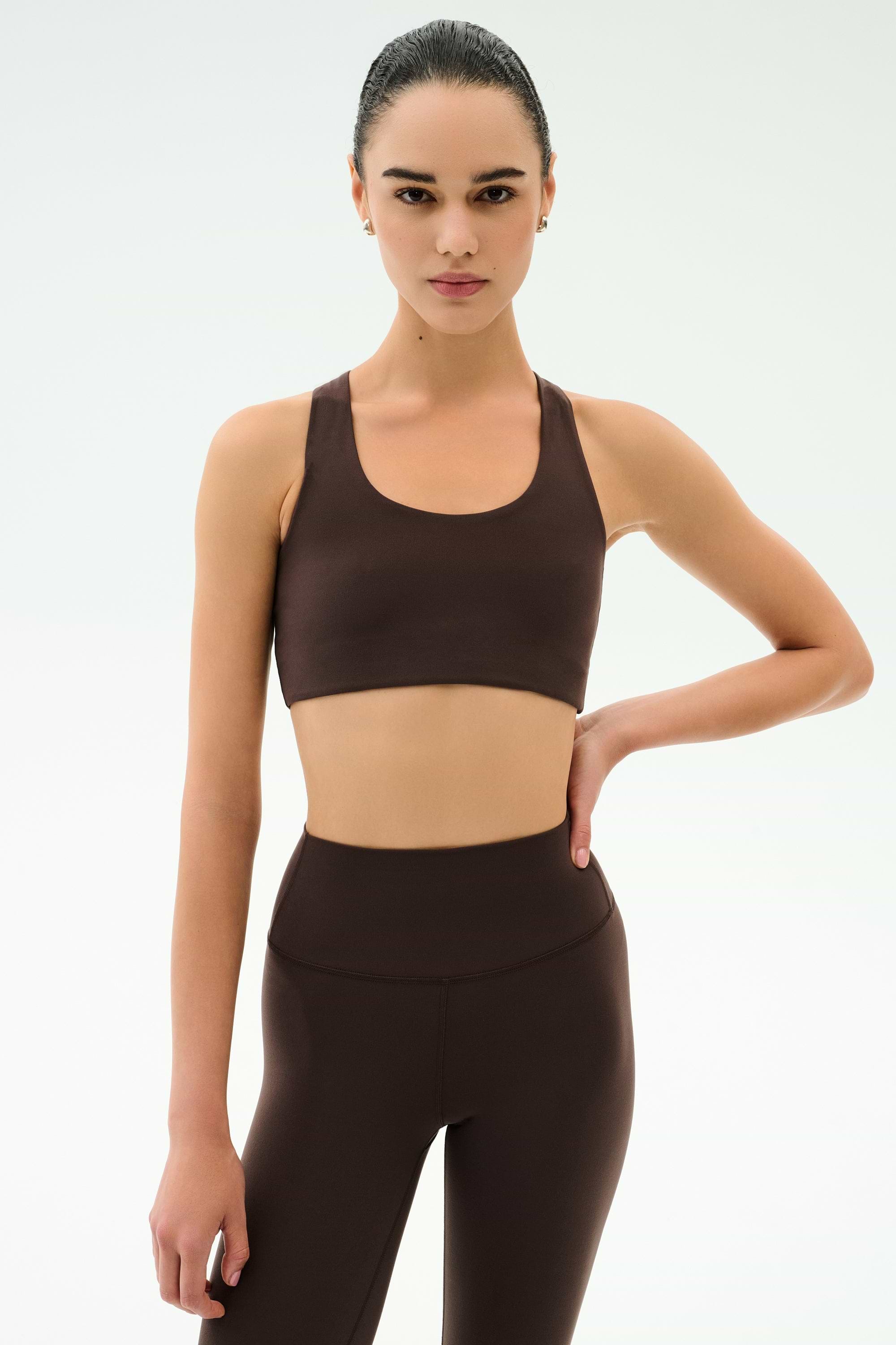 A woman poses against a plain background in SPLITS59’s Sara Airweight Bra - Dark Chocolate and matching leggings, one hand on her hip and a neutral expression—ideal for hot yoga or Pilates.