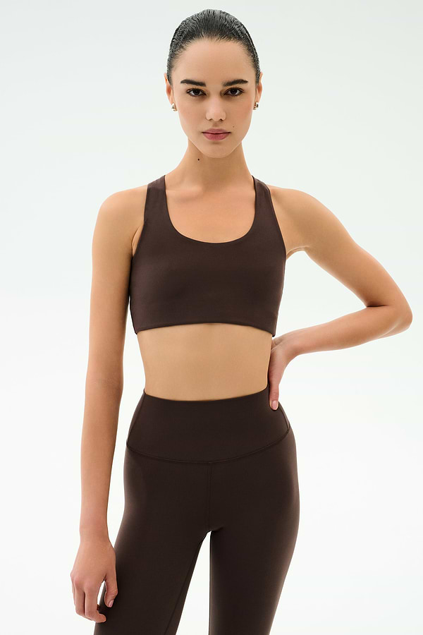 A woman poses against a plain background in SPLITS59’s Sara Airweight Bra - Dark Chocolate and matching leggings, one hand on her hip and a neutral expression—ideal for hot yoga or Pilates.