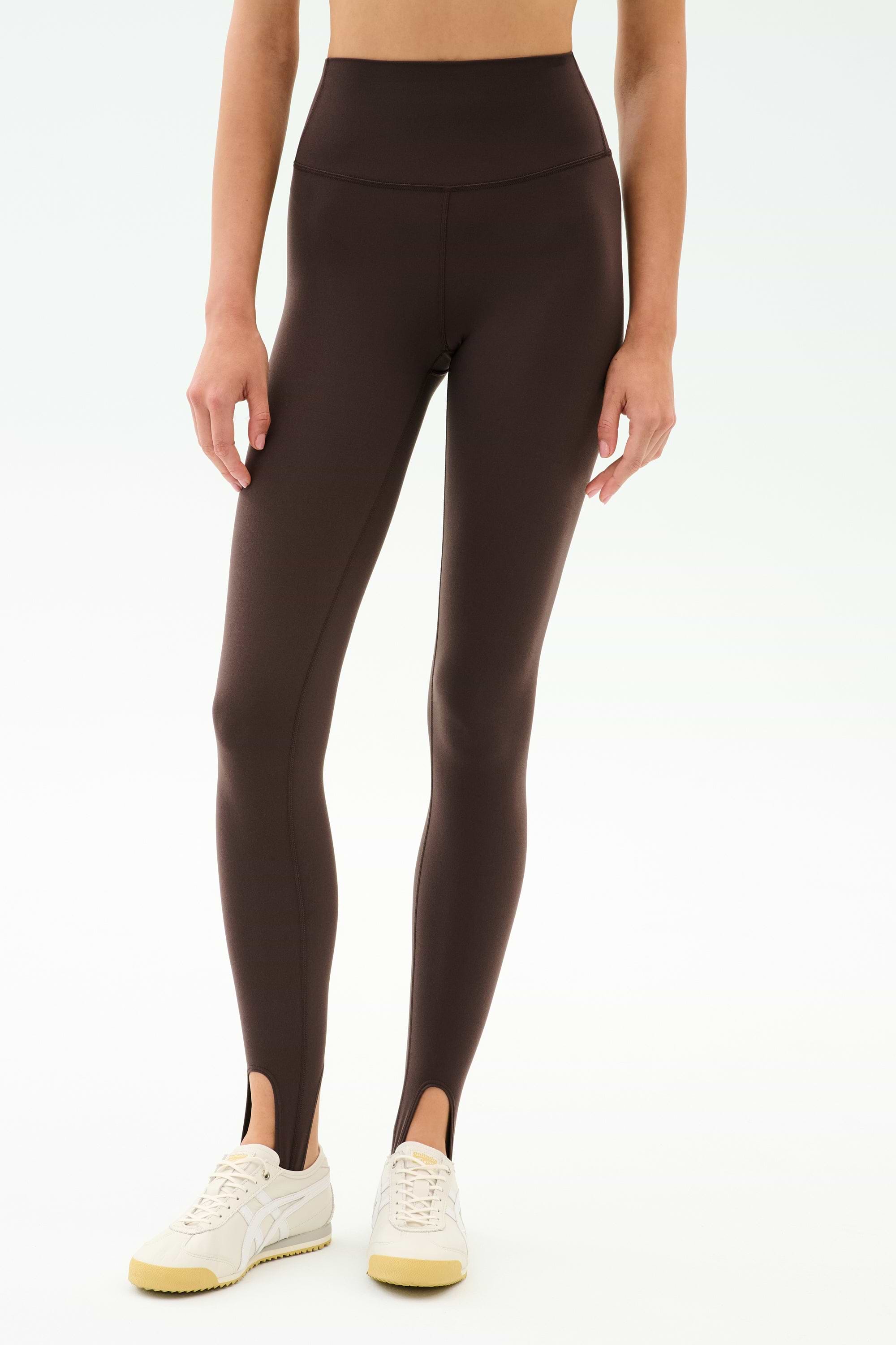 A person wearing SPLITS59 River High Waist Airweight Stirrup leggings in Dark Chocolate, paired with white sneakers, stands against a light background.