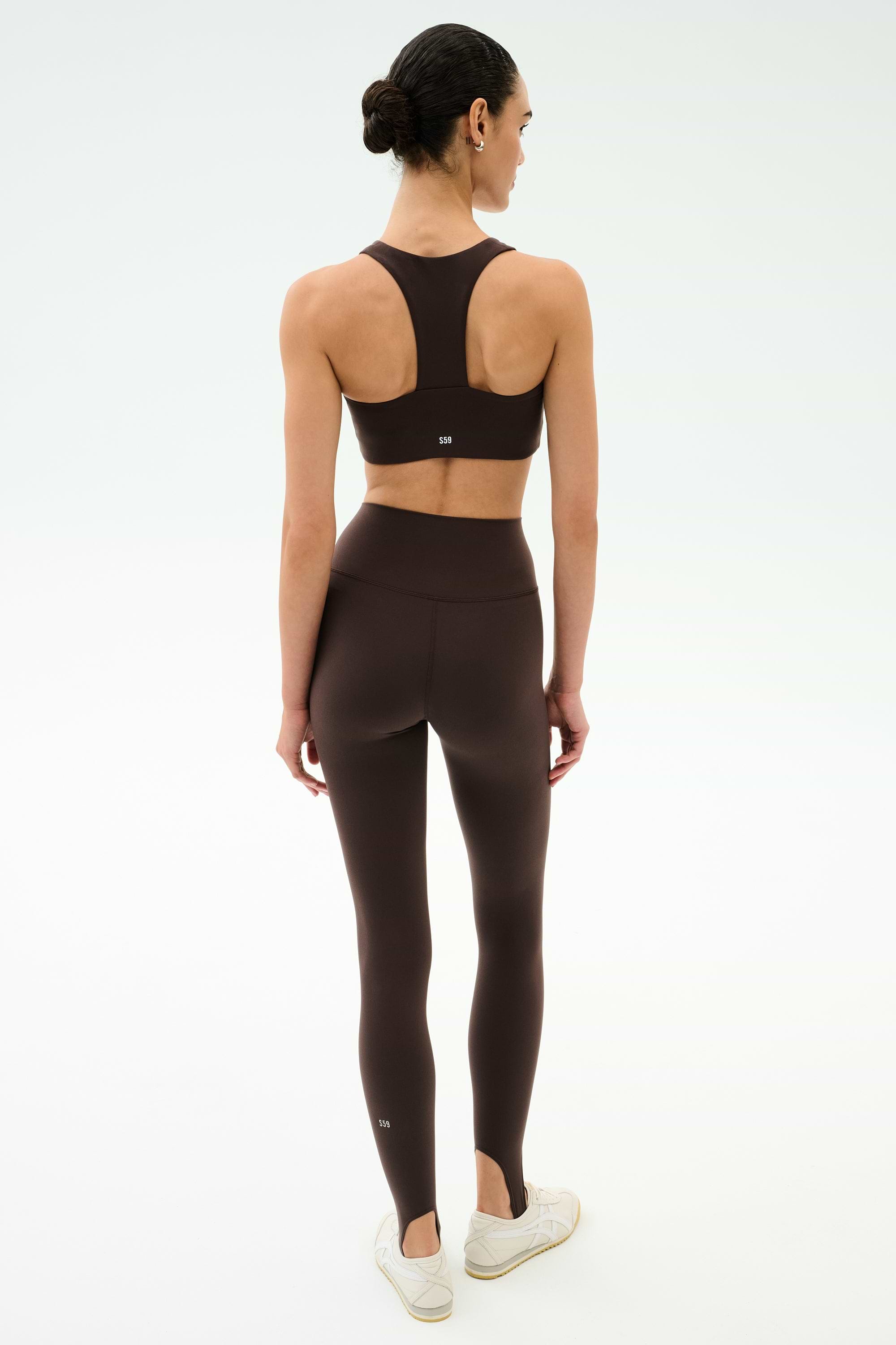 A woman faces away, wearing the SPLITS59 River High Waist Airweight Stirrup leggings in Dark Chocolate with a matching sports bra and white sneakers, set against a plain white background.