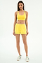 A woman stands against a white background, wearing SPLITS59's Ella Airweight Track Short in Bright Yellow/White with a yellow sports bra and white sneakers—ideal for hot yoga.