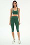 A woman faces forward in the SPLITS59 Ella High Waist Airweight Capri in Arugula/White, paired with a green sports bra and white sneakers, standing against a plain white background.