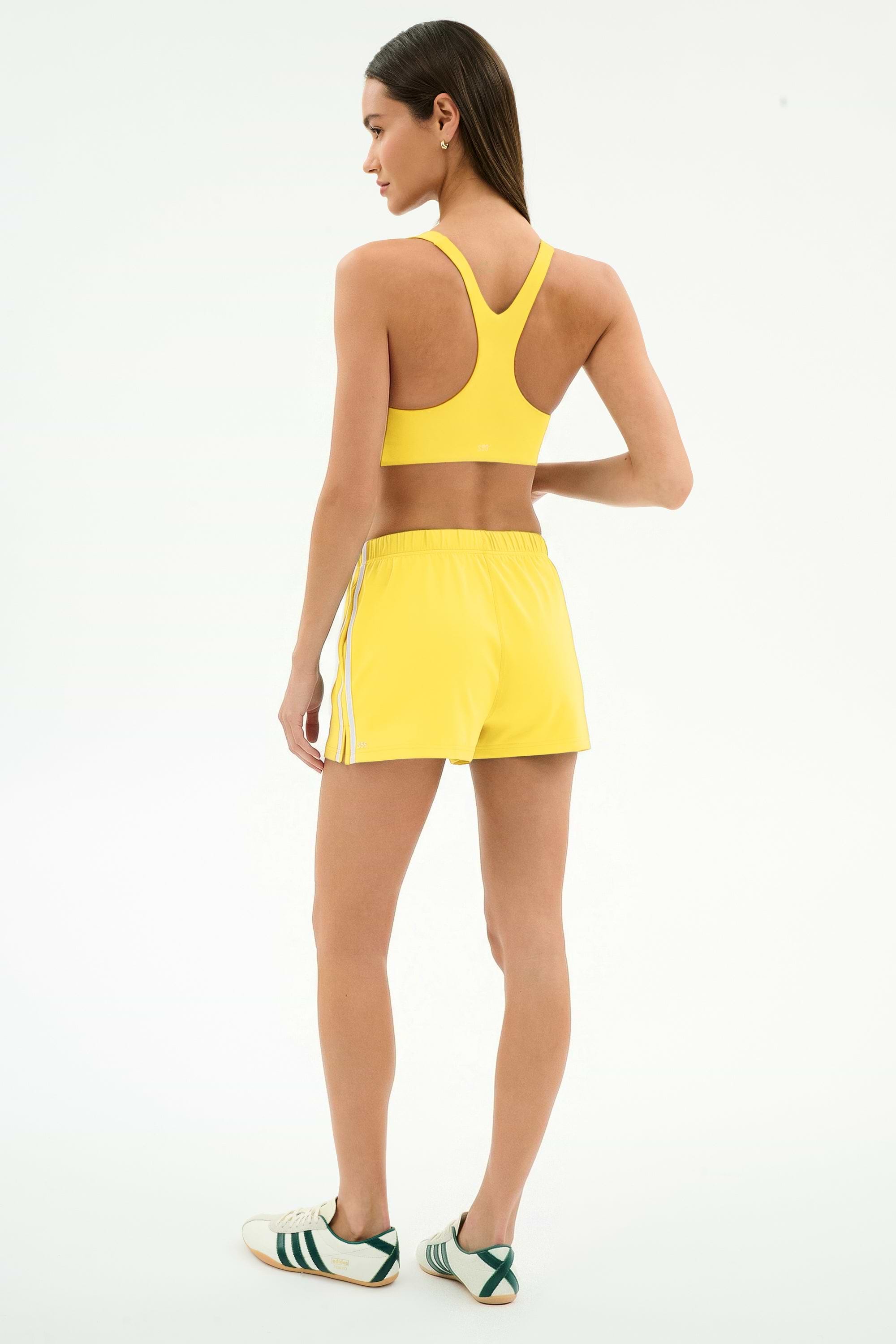 A woman stands facing away, wearing SPLITS59 Ella Airweight Track Short in Bright Yellow/White and a yellow sports bra, paired with white sneakers featuring green stripes.