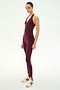 A woman stands in a studio wearing the SPLITS59 June Rib Halter in Dark Cherry—a matching burgundy halter top crafted from a soft modal spandex blend—paired with leggings featuring stirrups and beige Adidas sneakers.