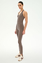 A person wearing the SPLITS59 June Rib Halter in Lentil, a taupe sleeveless fitted jumpsuit crafted from stretchy material and featuring stirrup leggings, styled with white sneakers and standing against a plain light background.