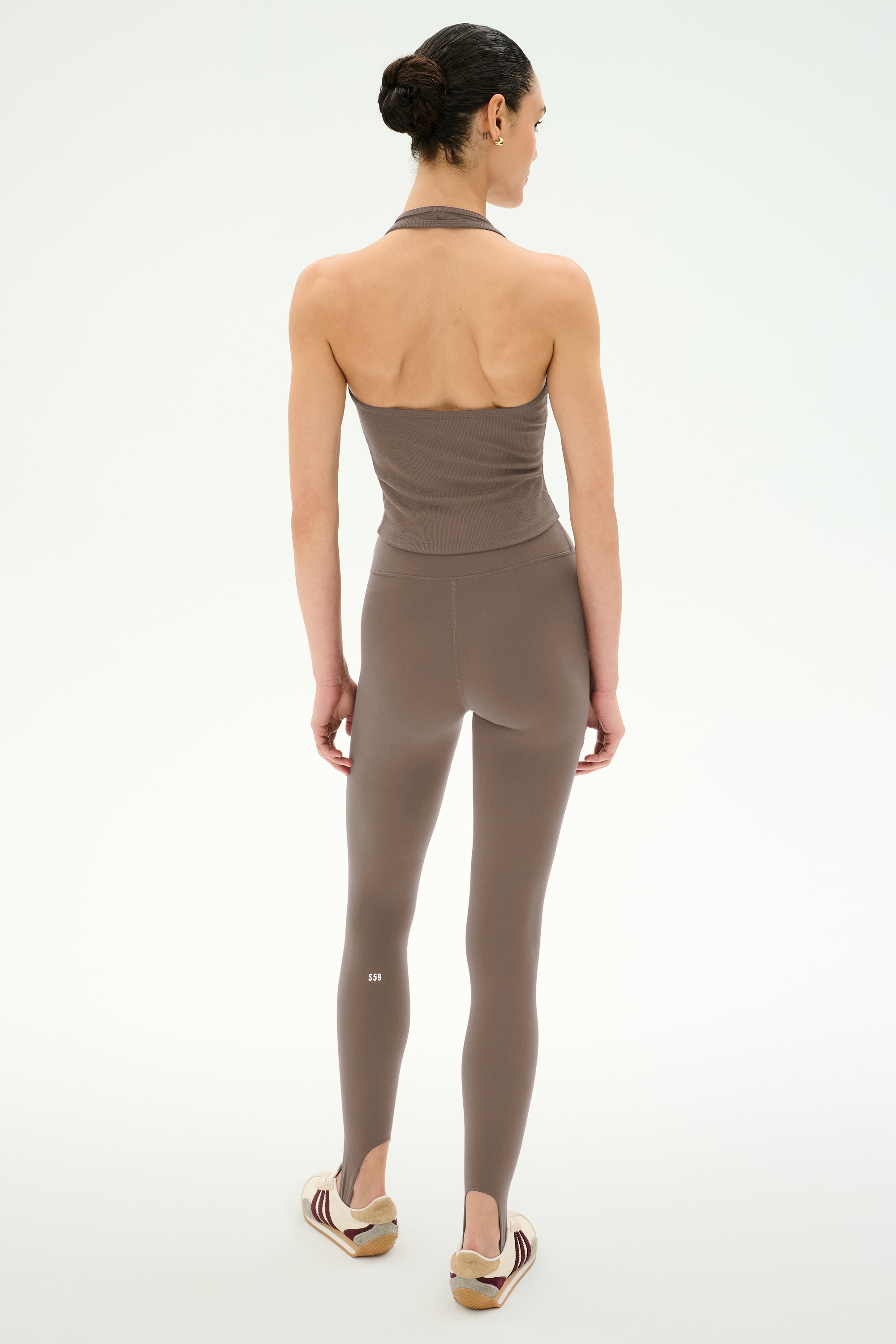 A woman stands against a white background, facing away, wearing the SPLITS59 June Rib Halter in Lentil paired with matching leggings, both crafted from stretchy material, and flat sandals.