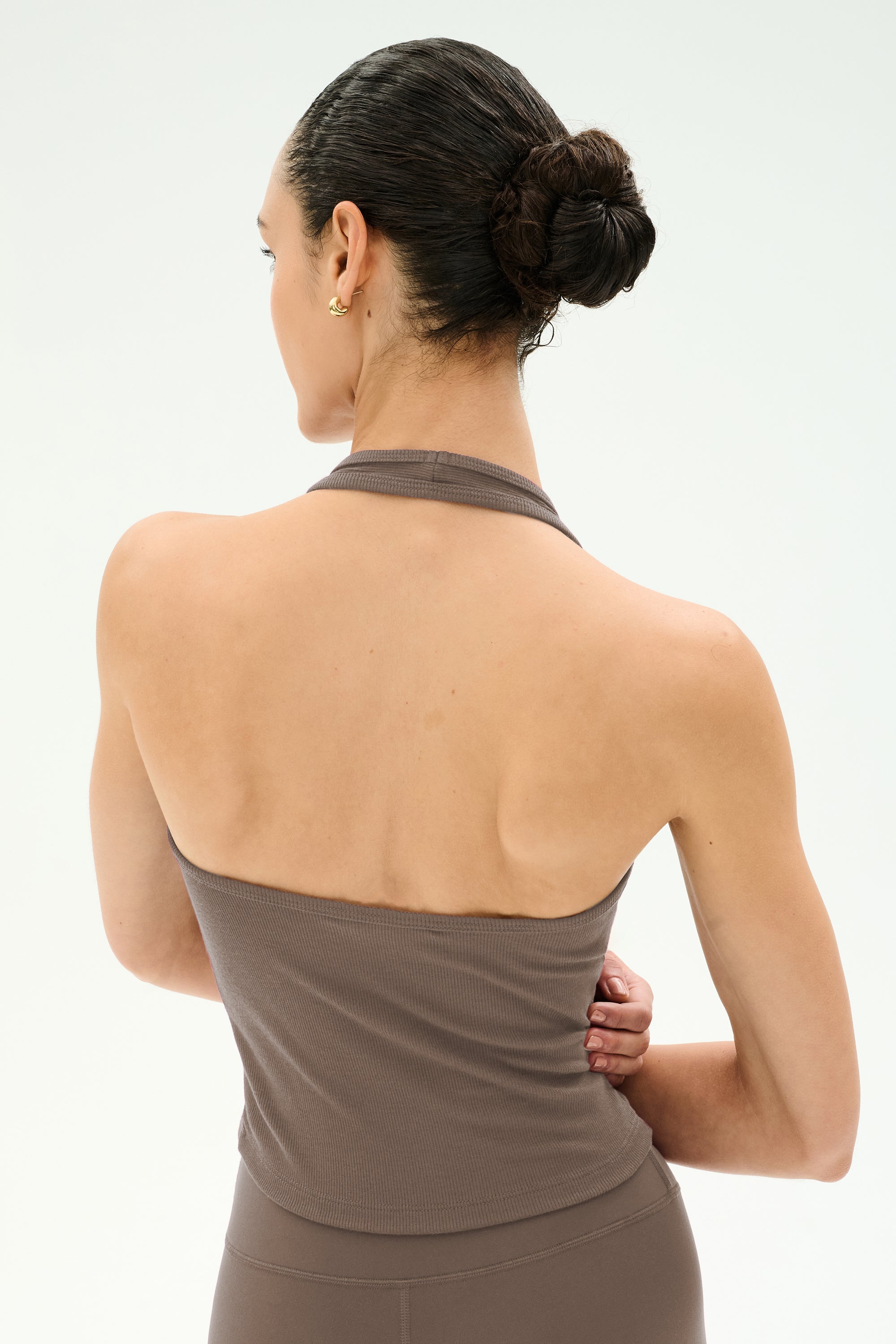 A woman with dark hair in a bun is seen from behind against a plain, light background, wearing the SPLITS59 June Rib Halter in Lentil—a taupe halter-style top crafted from soft modal fabric.