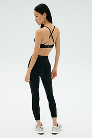 A woman wears the SPLITS59 Theo Rigor Bra - Black with black leggings and gray running shoes, standing and looking over her shoulder. Ideal for multi-sport performance, set against a plain background.