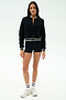 A woman faces forward against a plain white background wearing the SPLITS59 Willa Sherpa Jacket in Black, paired with black shorts, white socks with black stripes, and white sneakers.