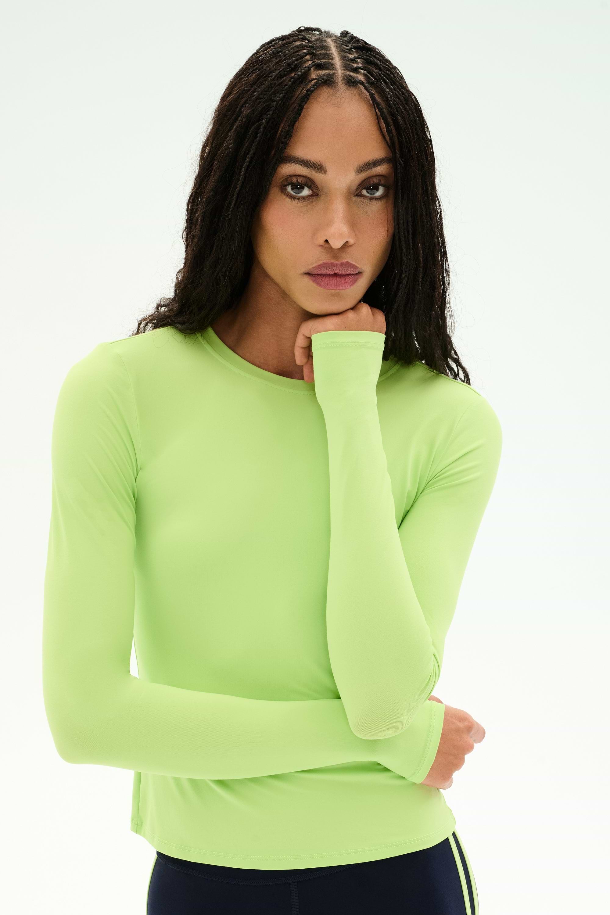 A woman with long braids wears the SPLITS59 Airweight Lite LS Waist Length top in Limeade—a fitted, long-sleeve style crafted from a soft nylon-spandex blend—paired with dark pants, as she rests her chin on her hand and looks forward against a plain light background.