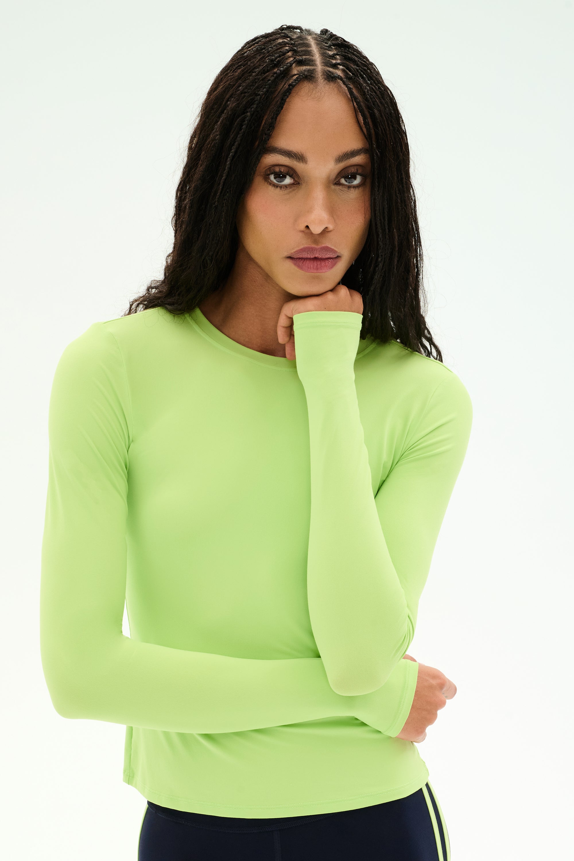 A woman with long braids wears the SPLITS59 Airweight Lite LS Waist Length top in Limeade—a fitted, long-sleeve style crafted from a soft nylon-spandex blend—paired with dark pants, as she rests her chin on her hand and looks forward against a plain light background.