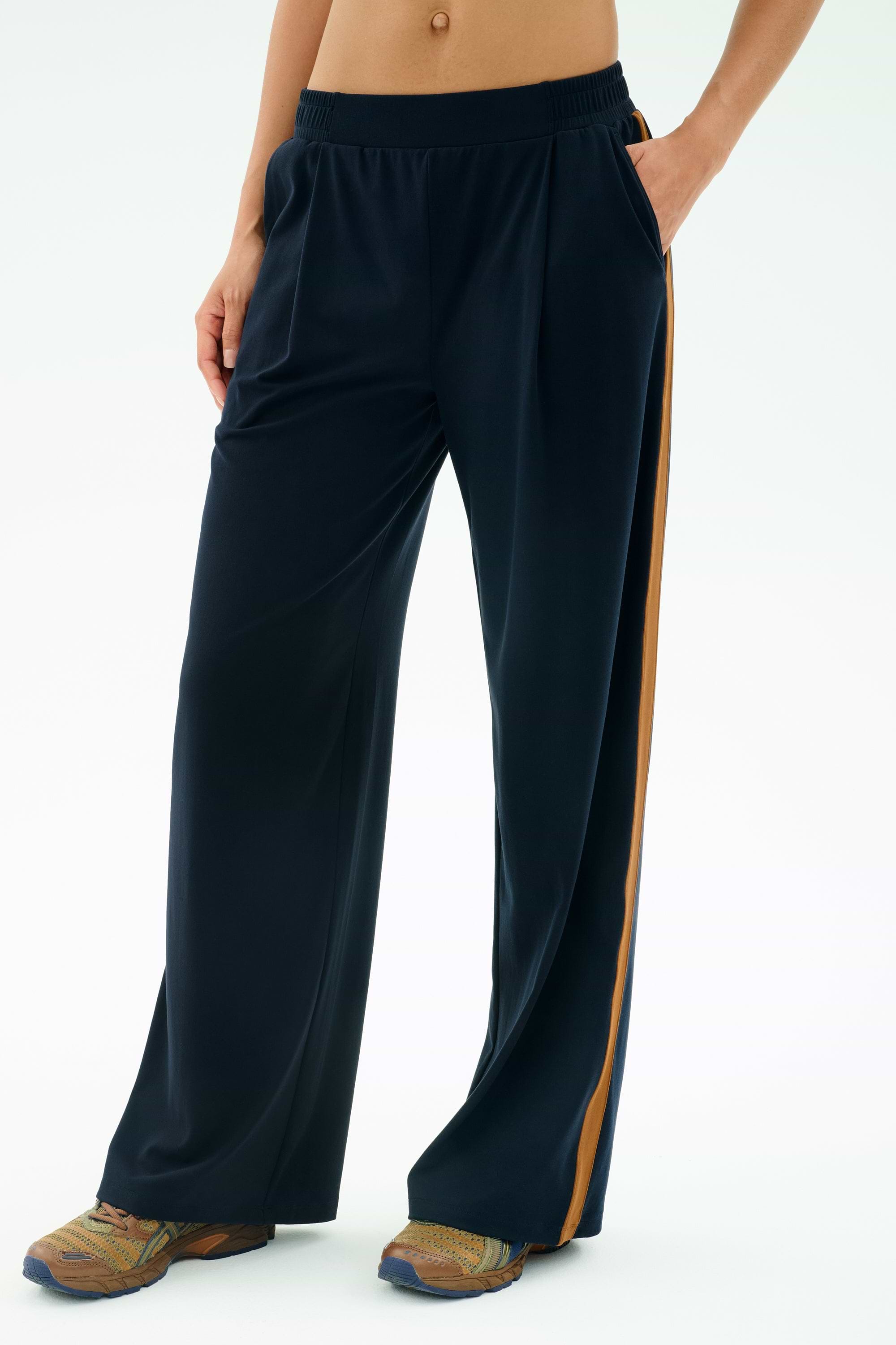 A person wears the SPLITS59 Luca Airweight Trouser with Stripe in Indigo/Honey, styled with colorful sneakers and a bare midriff, standing with one hand in their pocket against a plain background.