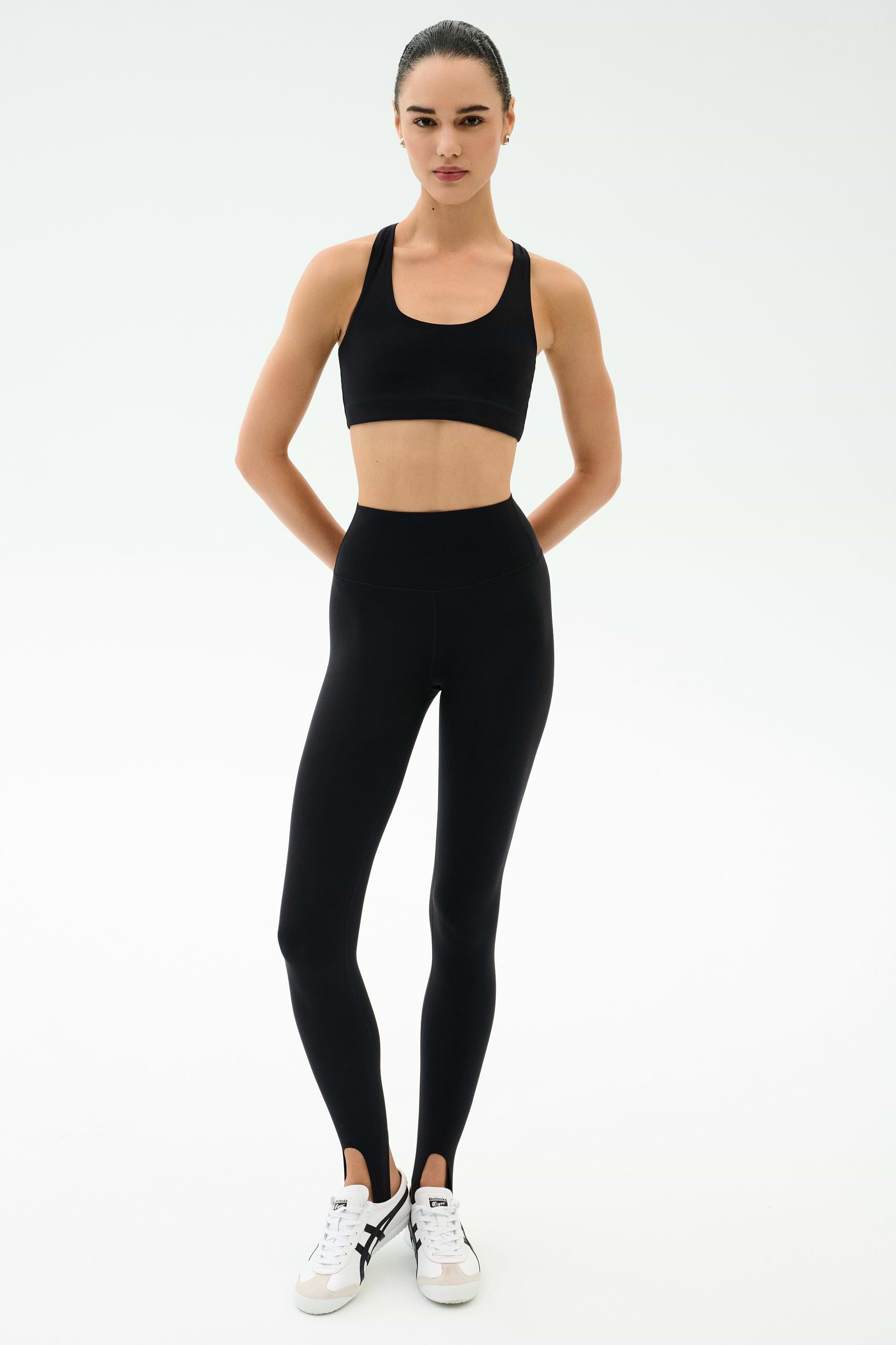 A person stands facing forward, wearing SPLITS59 River High Waist Airweight Stirrup leggings in black and a black sports bra, paired with white sneakers against a plain light background.