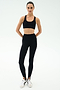 A person stands facing forward, wearing SPLITS59 River High Waist Airweight Stirrup leggings in black and a black sports bra, paired with white sneakers against a plain light background.