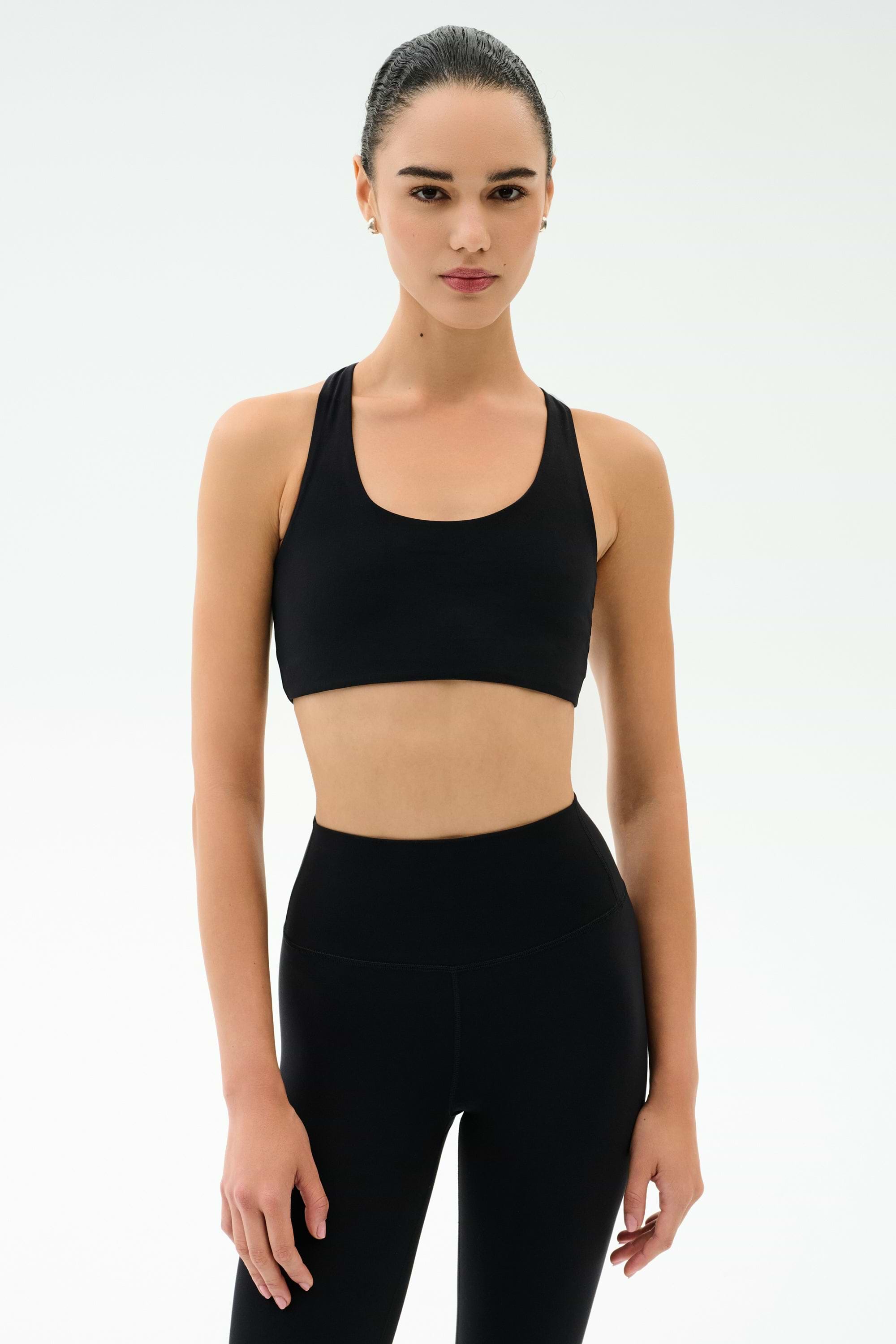 A woman faces forward in a black SPLITS59 Sara Airweight Bra and high-waisted leggings, ready for hot yoga or Pilates against a plain light background.