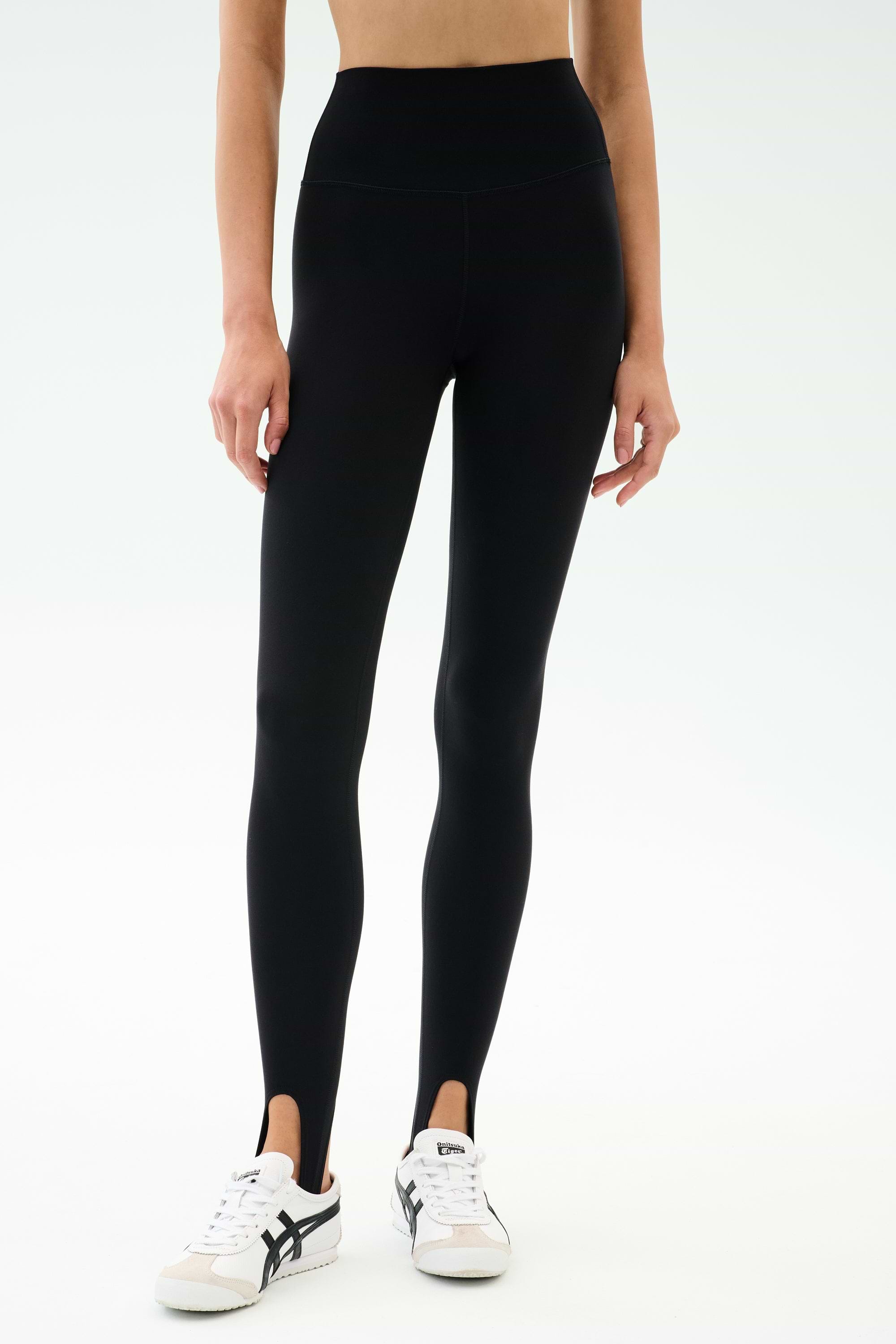A person wearing SPLITS59 River High Waist Airweight Stirrup leggings in black with white sneakers, standing against a plain light background.