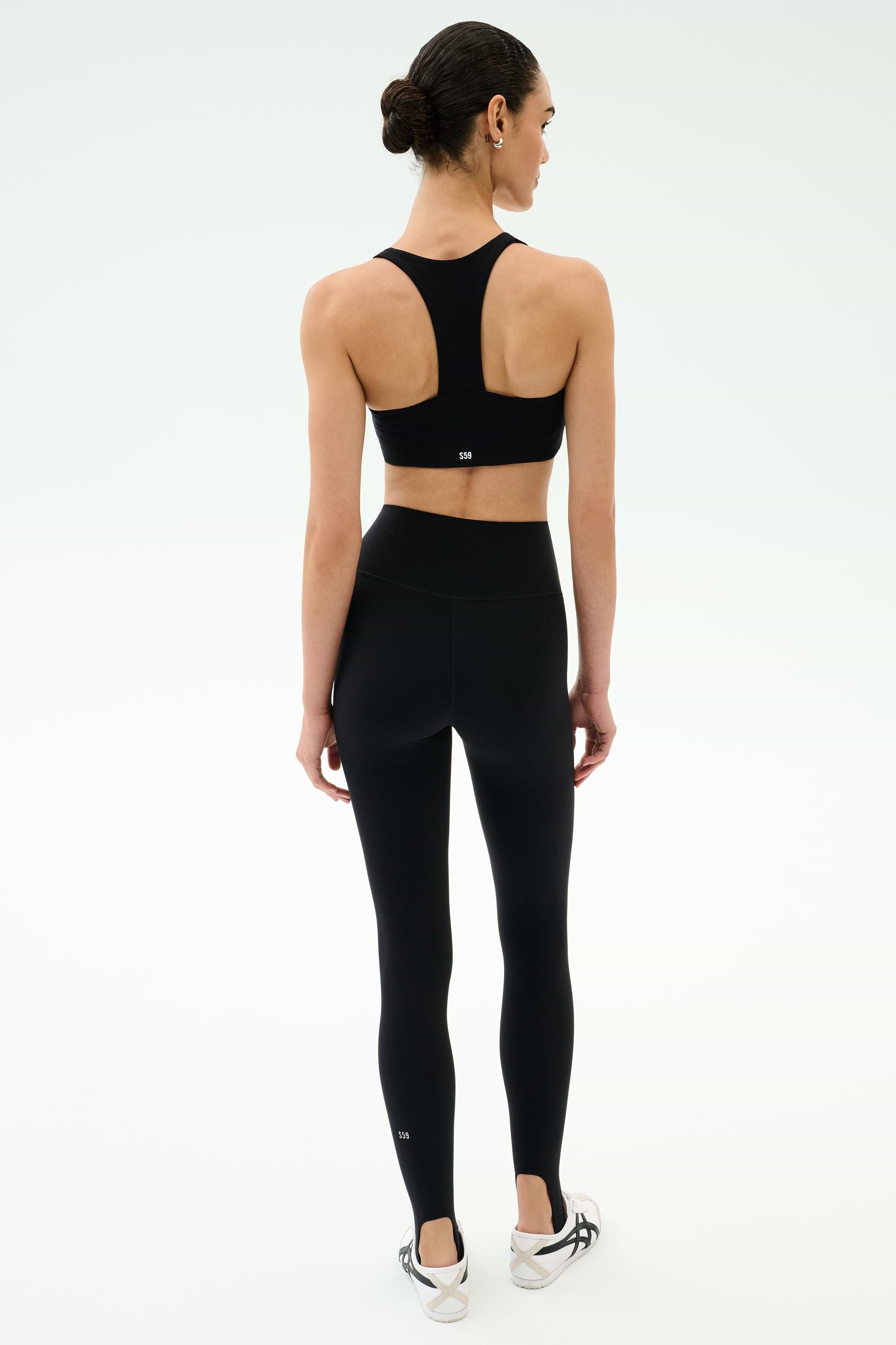 A woman faces away, wearing SPLITS59’s River High Waist Airweight Stirrup leggings in black with a matching sports bra and white sneakers, set against a plain white background.