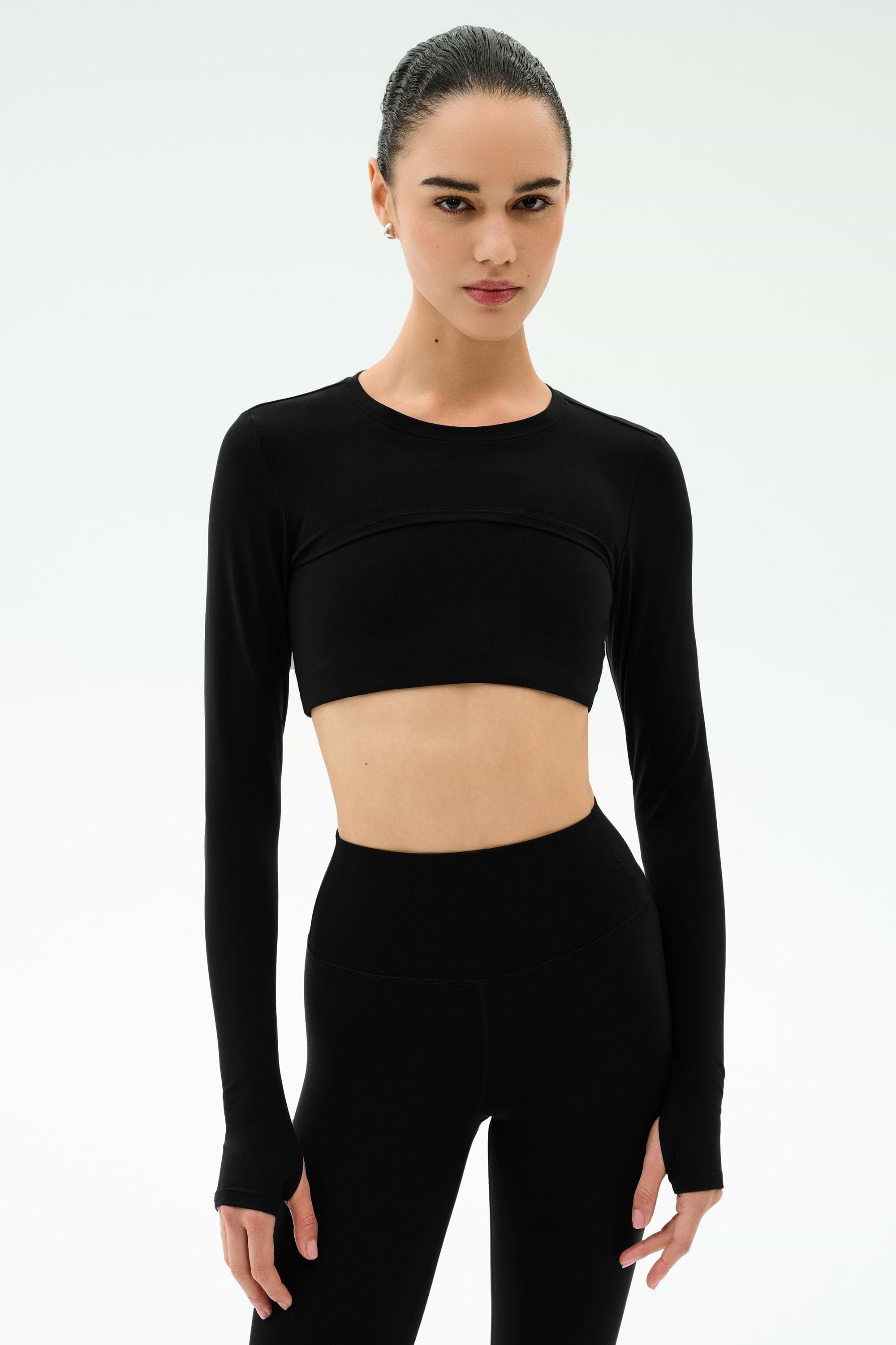 A woman stands against a plain white background wearing the SPLITS59 Amelia Airweight Lite Shrug in black with matching high-waisted leggings.