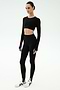 A person stands against a white background wearing the SPLITS59 Amelia Airweight Lite Shrug in black, paired with black long-sleeve crop top, black high-waisted stirrup leggings, and white athletic shoes.