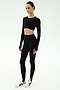 A person stands against a white background wearing the SPLITS59 Amelia Airweight Lite Shrug in black, paired with black long-sleeve crop top, black high-waisted stirrup leggings, and white athletic shoes.